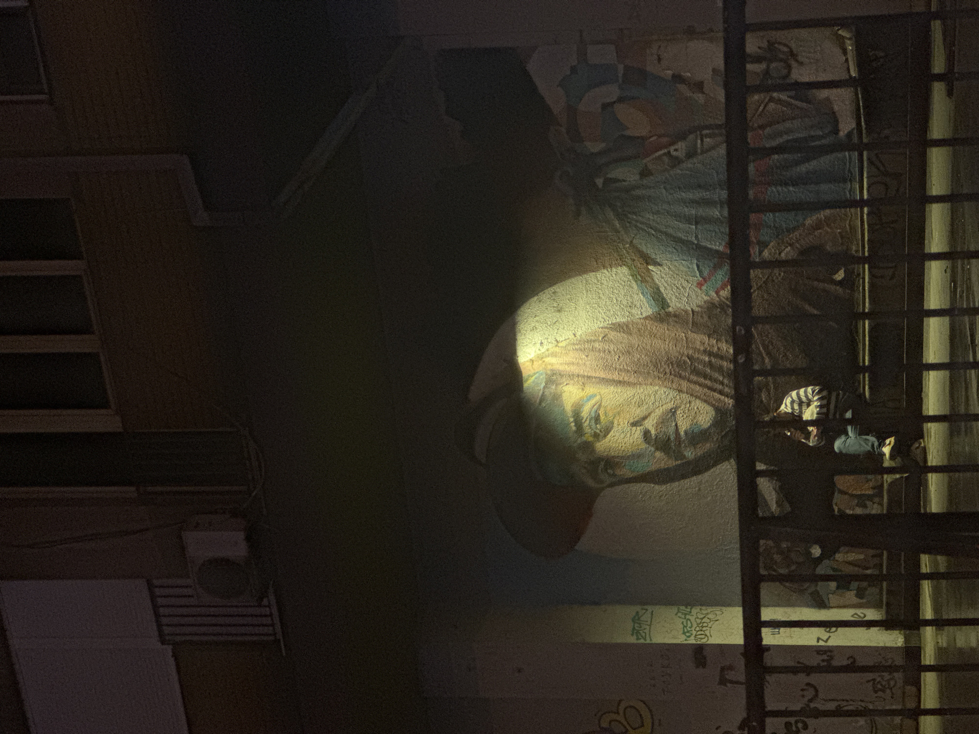 A person sits on a bench in front of a mural in Belgrade, Serbia, at night. The mural depicts a large face wearing a hat and a hooded garment, illuminated by a spotlight. The person is wearing a striped shirt and blue jeans, and is looking down at something in their hands. The scene is framed by a black metal fence. The wall also features other graffiti tags. An air conditioning unit is visible on the building above. The ground appears wet, suggesting it may have recently rained.