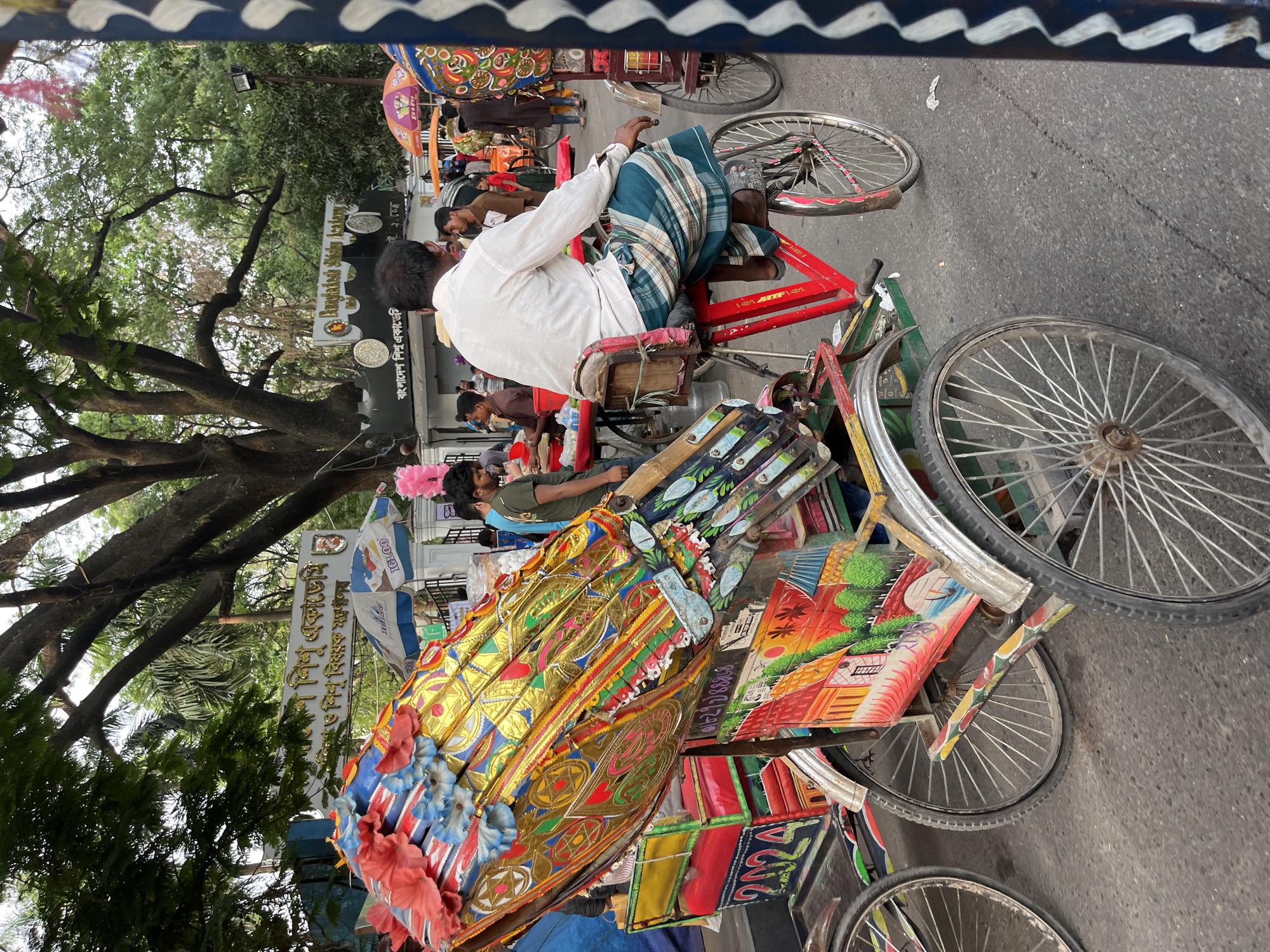 The image captures a street scene in Dhaka, Bangladesh, focusing on a rickshaw and its puller. The rickshaw is elaborately decorated with colorful paint, flowers, and metallic details, indicative of local artistry. The puller, dressed in a white shirt and a checkered lungi, sits on the vehicle ready to start, his back to the viewer. The street appears busy, with other people and rickshaws visible in the background, along with lush trees. The signpost reads "Bangladesh Shishu Academy", so this is near Bangladesh Shishu Academy.
