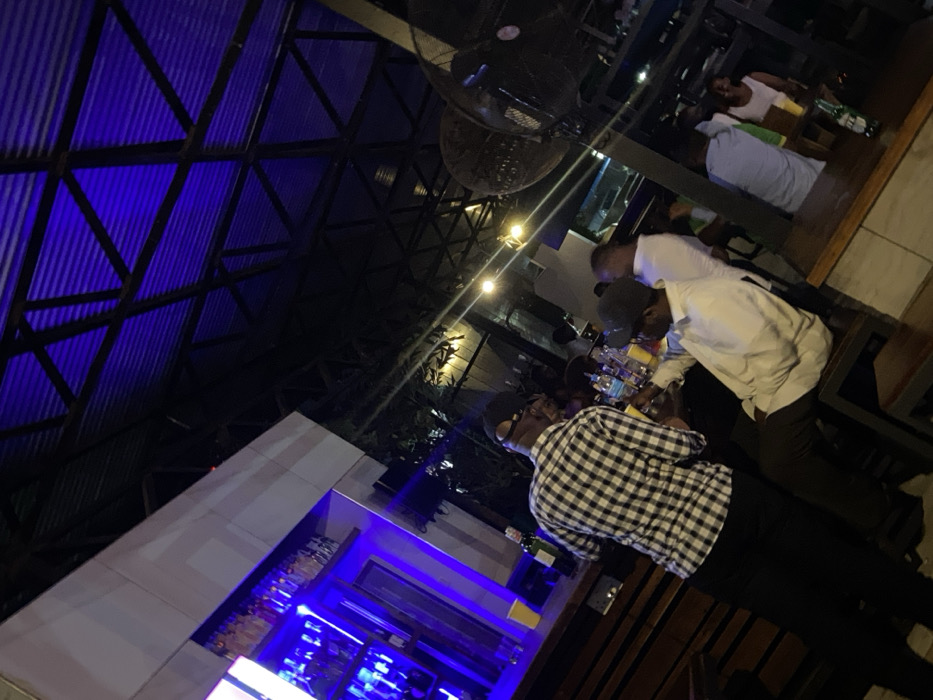 The image shows a bar in Dar es Salaam, Tanzania, located at the coordinates -6.727 latitude and 39.142 longitude. The bar is lit by a blue neon light, which reflects on the ceiling of the bar. There are several people in the bar, including a man wearing a black and white plaid shirt and a man wearing a white shirt. Both men are holding yellow cups, possibly containing a drink. There is a long wooden bar to the left of the image and shelves on the right side. There is a large fan in the corner of the bar and many more people are visible in the background. The bar has a modern feel and the environment is relaxed and casual.