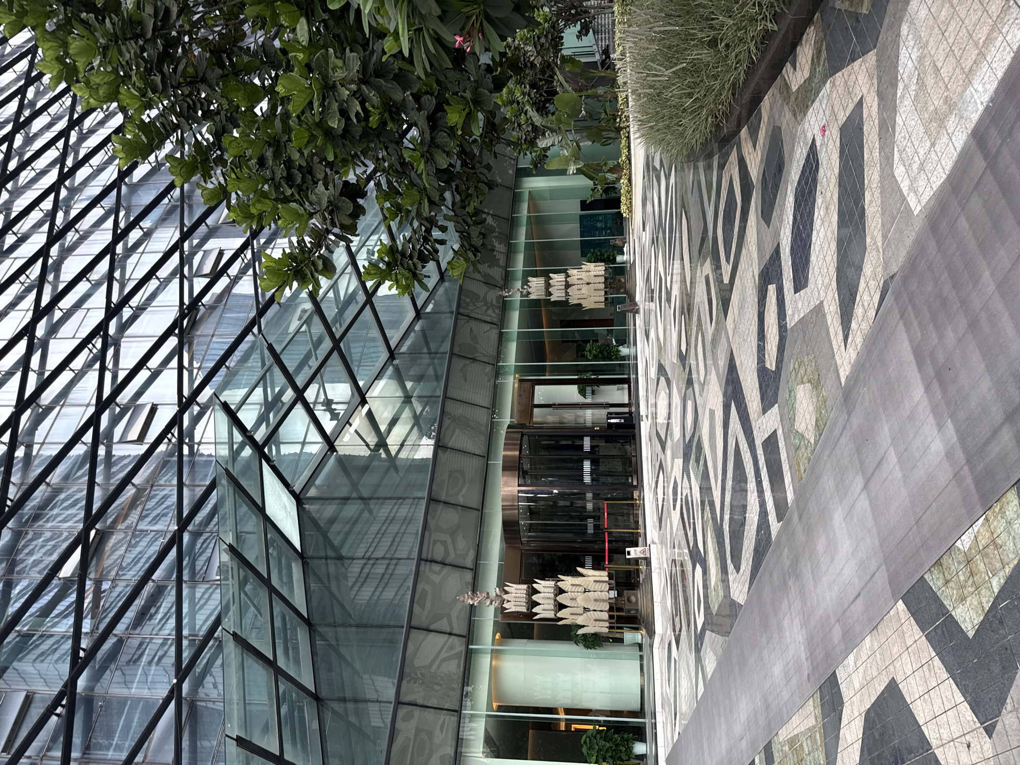 The image depicts a modern architectural building with a glass facade and a geometric patterned outdoor pavement. Green foliage from trees is visible in the upper left portion of the frame, casting shadows on the glass structure. The building's exterior is characterized by a lattice of black metal beams supporting large glass panels, creating a reflective and somewhat abstract surface.
In the foreground, the pavement features a repeating pattern of interconnected geometric shapes in shades of gray, black, and white. A dark gray walkway runs diagonally across this patterned area. Beyond the glass facade, a revolving door entrance to the building is visible. Inside, the interior appears to be a well-lit lobby or atrium, with modern lighting fixtures and some potted plants. A piece of modern art, consisting of stacked, layered elements, is suspended in front of the entrance.
The scene appears to be an outdoor space in a contemporary urban environment, likely during daylight hours given the lighting. The overall impression is one of sleek, modern design and a focus on natural light and geometric aesthetics. There are no people clearly visible in the frame, suggesting an unoccupied moment or a focus on the architecture and design elements. There is no visible text in the image.