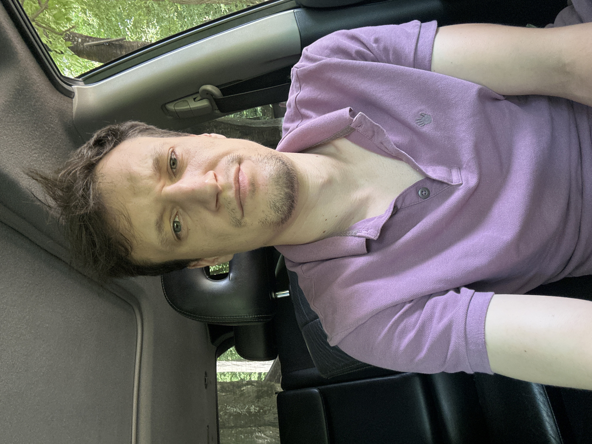 A man is seated in the driver's seat of a vehicle. He is wearing a light purple polo shirt. The interior of the car is visible, including black leather seats and the headliner. The sunroof is open, allowing natural light to illuminate the scene. Through the open sunroof and the open driver's side window, trees and foliage are visible, suggesting an outdoor environment. The man appears to be looking directly at the camera with a neutral expression. The scene is captured from a frontal perspective. The location in the city of Humentsi, Ukraine cannot be determined from the provided image.
