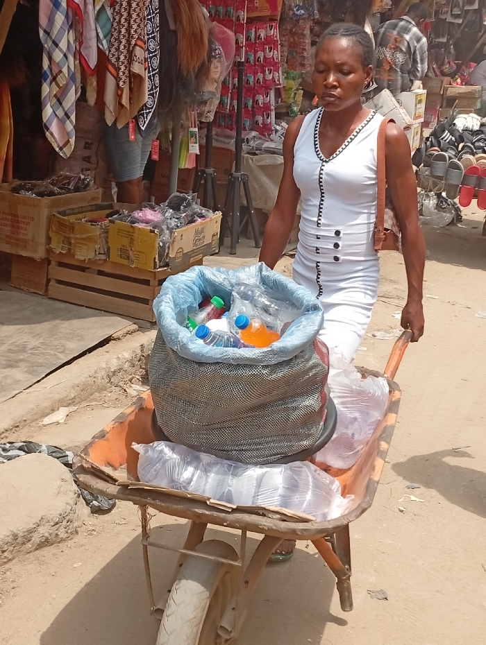 An adult female, appearing to be of African descent, is depicted outdoors in Fegge, Nigeria, actively pushing an orange-colored, single-wheeled wheelbarrow on an unpaved dirt surface. The woman has short, braided hair and wears a sleeveless white dress with black trim around the V-neck and black decorative buttons down the front. A light brown shoulder bag hangs from her left shoulder. The wheelbarrow's tray contains a large, grey mesh bag with a blue plastic lining filled with various plastic bottles of beverages, including water and orange-colored liquid. Several clear plastic bags containing other packaged items are also present in the wheelbarrow. In the background, to the left, market stalls display hanging scarves, wigs, and stacked cardboard boxes filled with merchandise. To the right, another stall exhibits numerous pairs of sandals on racks. The scene is brightly lit by daylight, casting shadows.