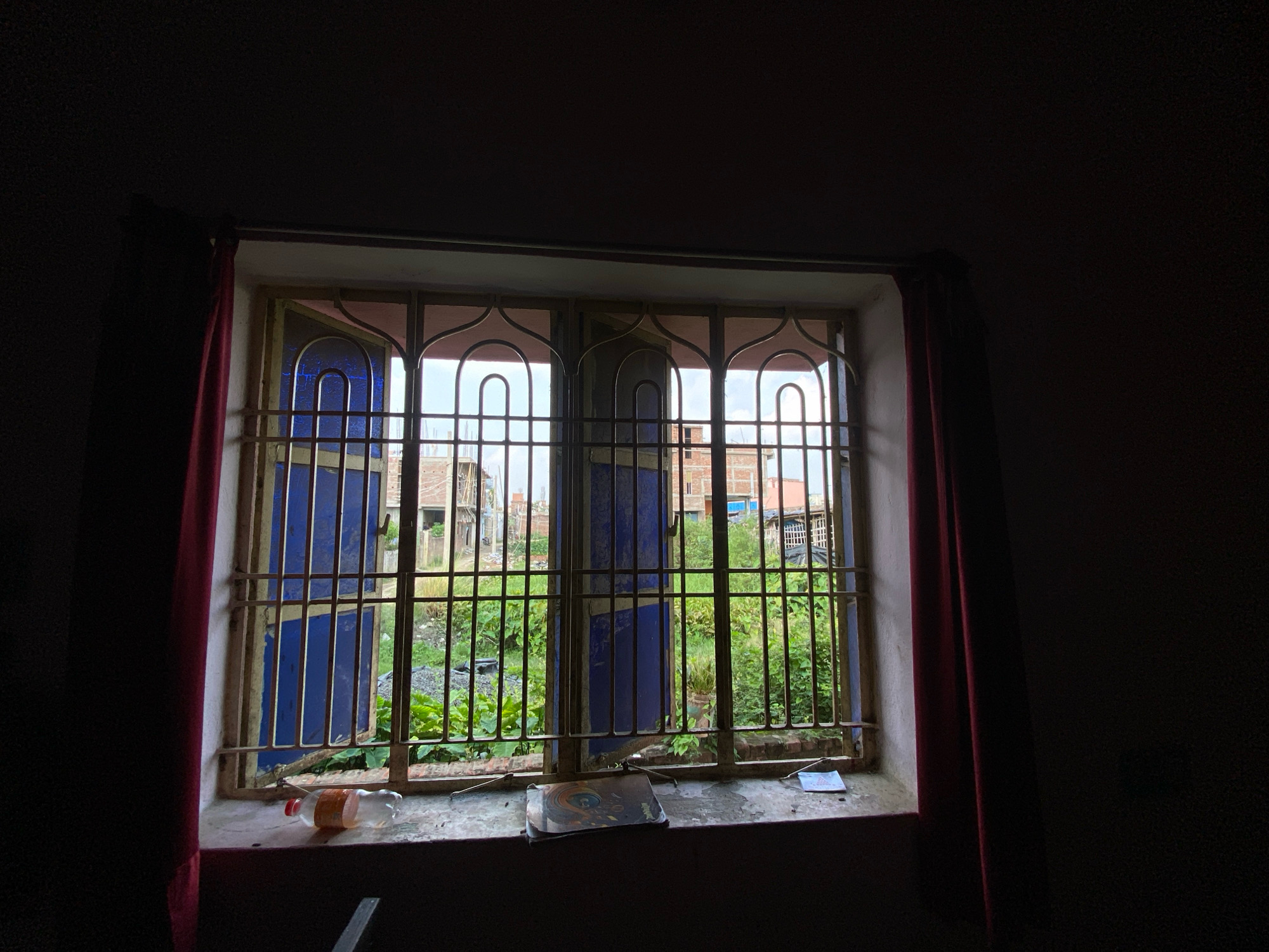 A medium shot captures an interior view through a barred window looking outwards onto a green, overgrown area with buildings under construction in the background. The window frame is painted light pink and has decorative metalwork. A metal security grate with vertical bars and curved decorative elements covers the window. Behind the grate, blue panels are visible. To the left and right of the window are dark red curtains. The windowsill, located in the foreground and darker than the window area, holds a partially filled plastic bottle with a red cap, a stack of what appear to be discs or flat objects, and a small white card. The external environment appears to be a rural or semi-urban setting in Bahadurpur, India, with evidence of ongoing construction, including exposed brickwork and scaffolding on multiple buildings. The vegetation outside is dense and green, suggesting a tropical or sub-tropical climate. The overall lighting suggests it is daytime, but the interior is dimly lit, creating a contrast with the brighter exterior.
