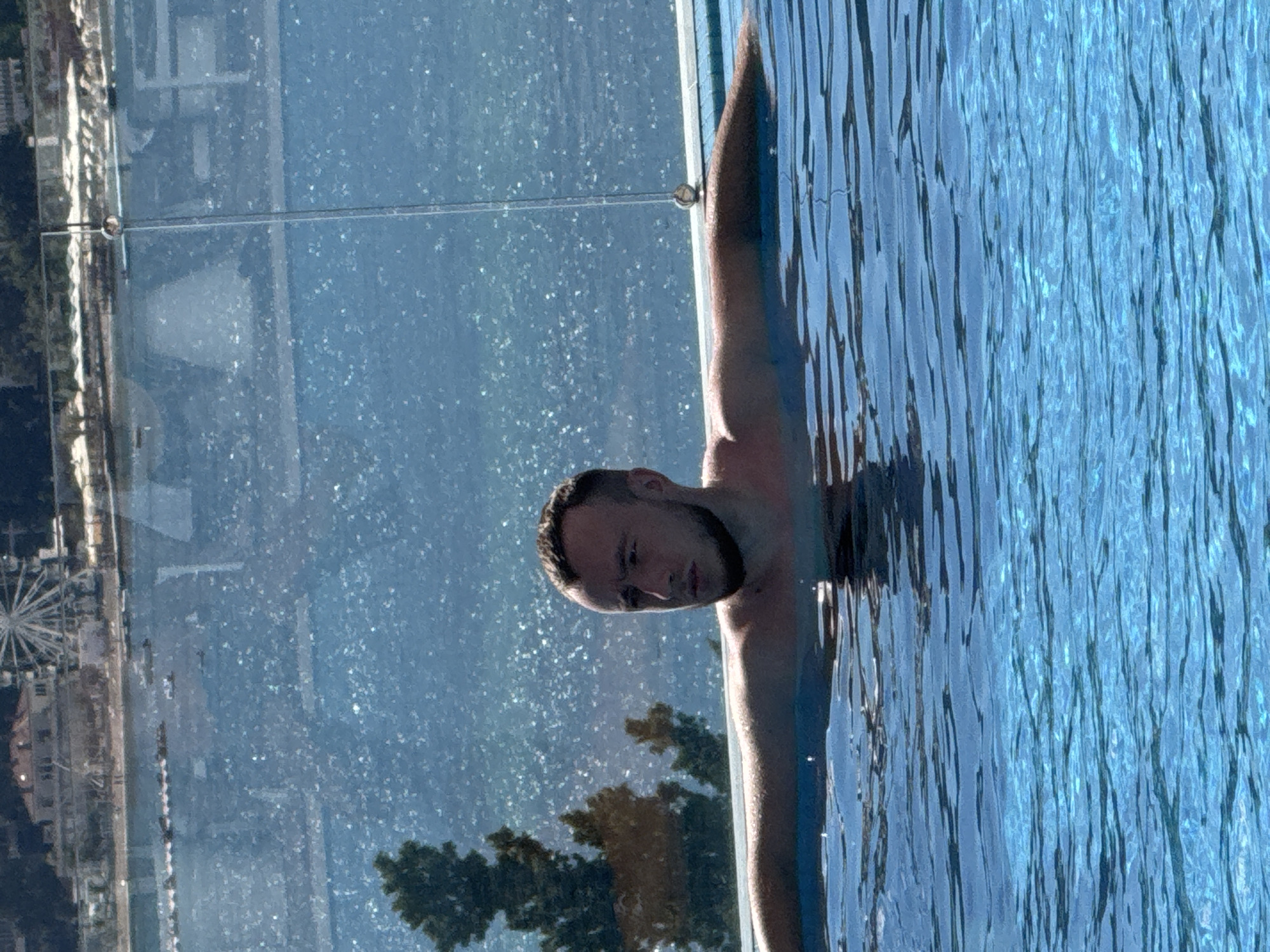 A man is in an infinity pool in Bečići, Montenegro, with a view of the Adriatic Sea and the Sveti Stefan island. The man is looking to his right, with his arms resting on the edge of the pool. The water is blue and clear, and the sky is also blue. In the background, there is a Ferris wheel and a resort. The image is taken from a slightly elevated angle.