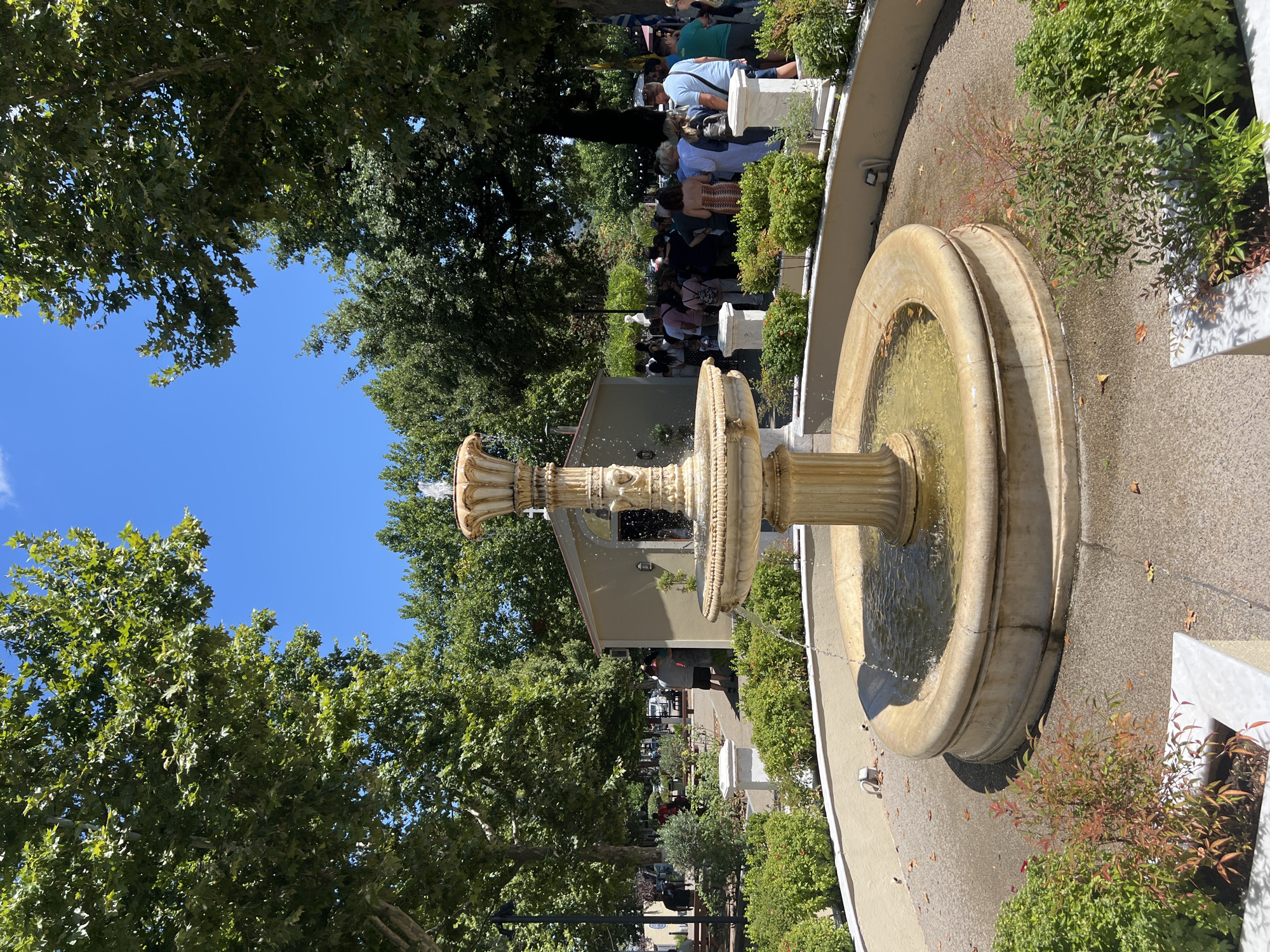 The image depicts a public space with a multi-tiered fountain as the central subject. The fountain, made of light-colored stone, features water cascading from its upper basin into the lower one, which is filled with water. Around the fountain, there is a circular pathway and manicured green bushes. In the background, several large trees with dense green foliage provide shade. Beyond the trees, a light-colored building with a pointed roof is visible. A crowd of people is gathered in front of the building, many appearing to be standing in a line or conversing. The sky is clear and blue, indicating a sunny day. The scene appears to be a plaza or a park area in the town of Prokopi, Greece, given the context of the provided information.