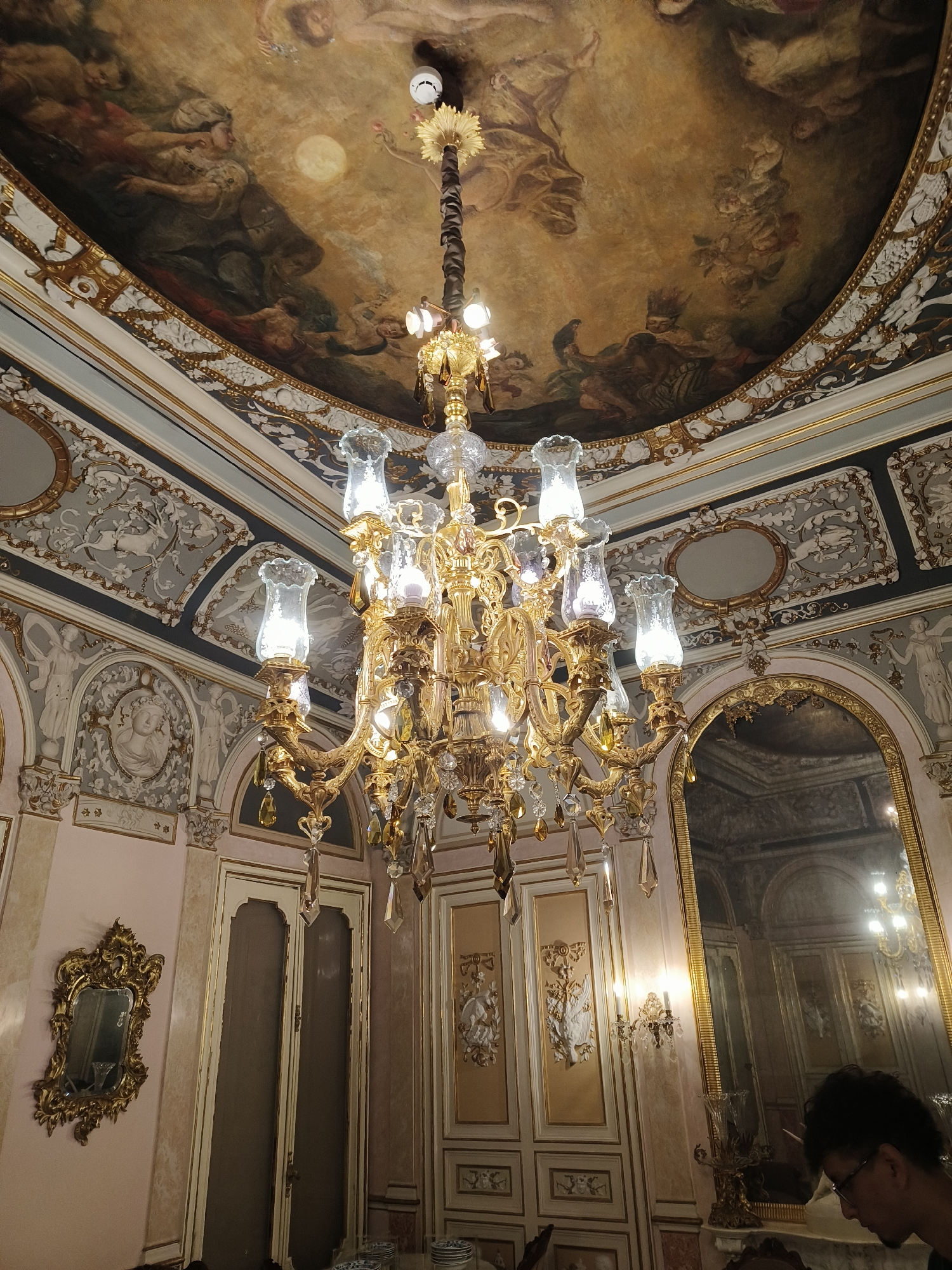 The image depicts the interior of a room with opulent Baroque-style decor. A large, ornate gold chandelier with multiple lights and crystal embellishments hangs from the center of the ceiling. The ceiling itself is adorned with a fresco depicting figures and celestial bodies, characteristic of grand historical buildings. The walls are elaborately decorated with moldings, bas-relief sculptures of figures and mythological scenes, and framed panels. A large, gilded mirror with an arched top reflects the opposite side of the room, showcasing more of the same elaborate detailing and a matching chandelier. To the left, a small, heavily ornamented gold mirror hangs on a pink-toned wall above a pair of tall, cream-colored doors with ornate gold trim. To the right, a person wearing glasses and a dark shirt is partially visible, looking down. The room appears to be a formal dining or reception area. This scene is located at the Palau de la Música de Valencia, Spain.