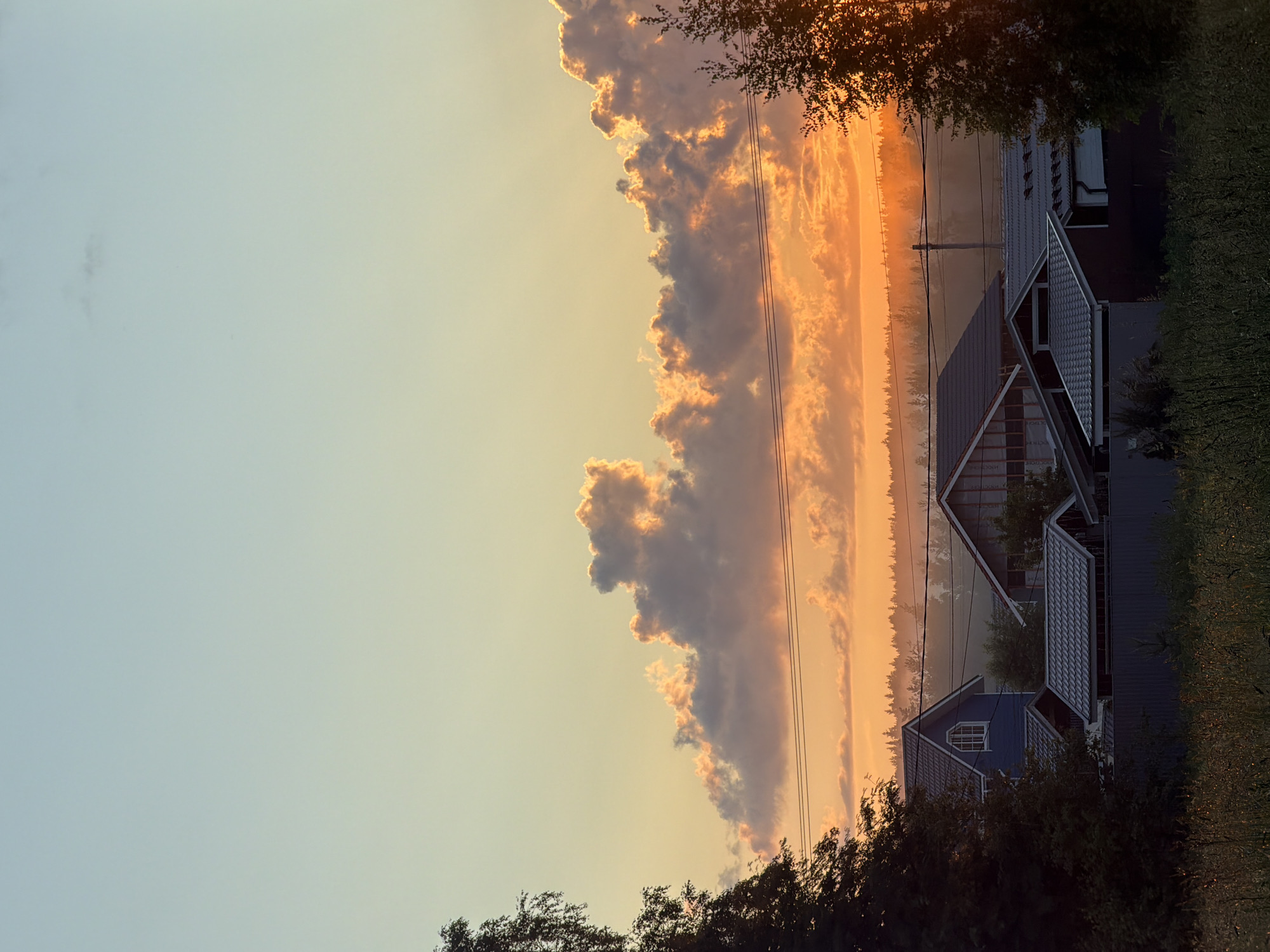 The image depicts a rural or suburban landscape at sunset. The sky is a gradient of soft blues, yellows, and oranges, with dramatic, illuminated clouds. In the foreground, the tops of several houses with gray roofs are visible, nestled among trees. A dark fence runs horizontally across the middle ground, separating the houses from a field of tall grass in the foreground. Electrical wires are strung across the scene, spanning between utility poles. The overall impression is one of a tranquil evening in a residential area. This scene is not located in the city of Mars, Russia, as Mars is a planet, not a city in Russia.