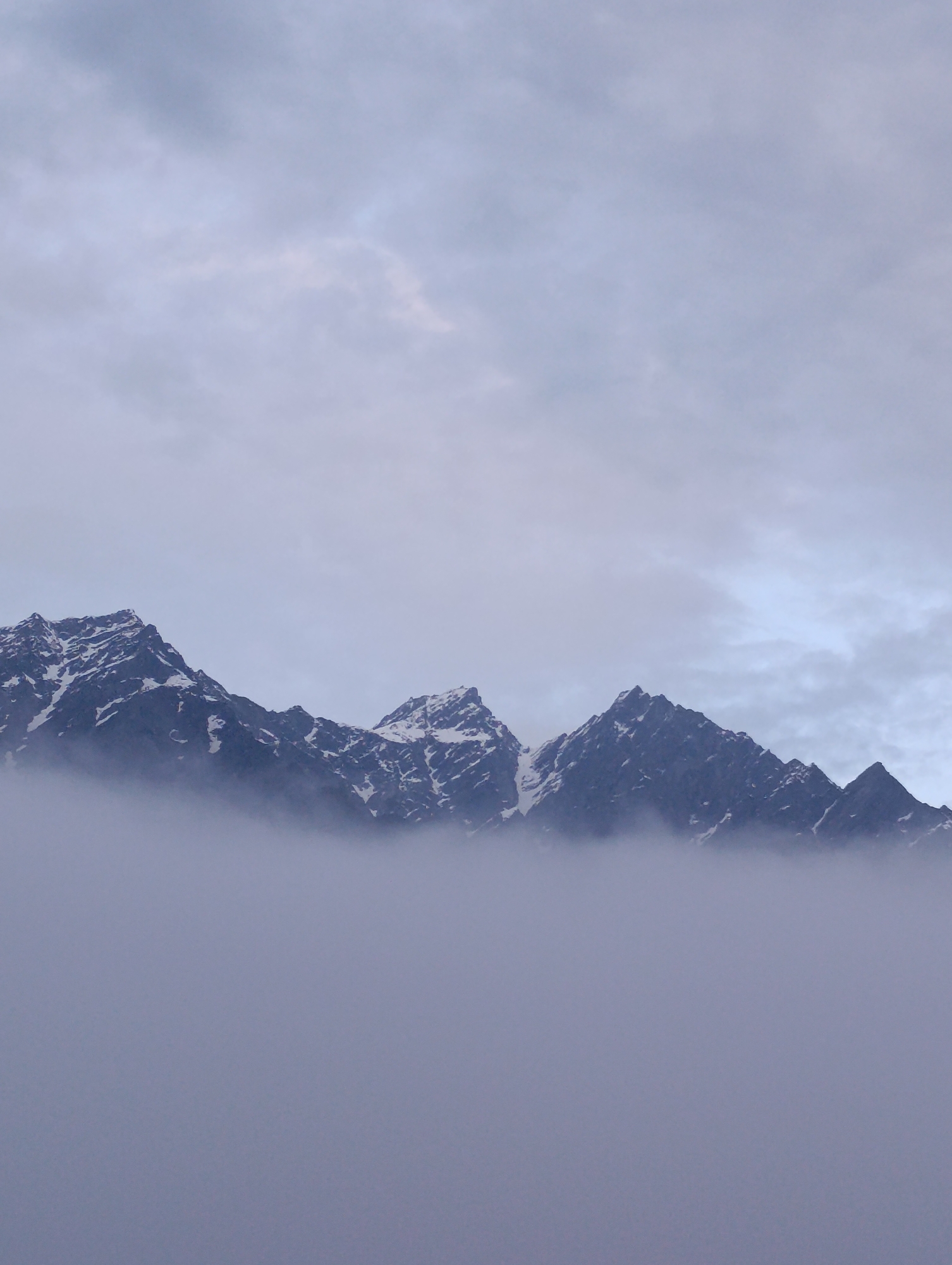 The image shows a dramatic mountain range partially obscured by thick, ethereal fog. The peaks are jagged and appear to be snow-capped, with dark, rocky slopes contrasting with patches of white snow. The sky above is overcast with muted tones of grey and white clouds. The fog blankets the lower portions of the mountains, creating a sense of depth and mystery. There are no people or man-made objects visible in the scene. The environment is natural and appears to be a remote, high-altitude location. While the image depicts mountains, there is no direct information to confirm it is specifically in Vashisht, India.