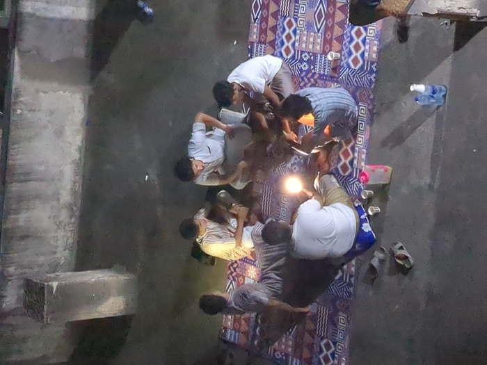Six men are sitting on a colorful patterned blanket spread out on a concrete surface, illuminated by a lamp. The men are all wearing casual clothing and appear to be conversing. Two empty plastic cups and a pink water bottle are visible on the blanket. Two bottles of water are on the concrete floor, to the right of the blanket. The location is a rooftop in Dar es Salaam, Tanzania. The concrete surface appears to be a rooftop or a balcony. The setting sun casts a warm glow on the scene.