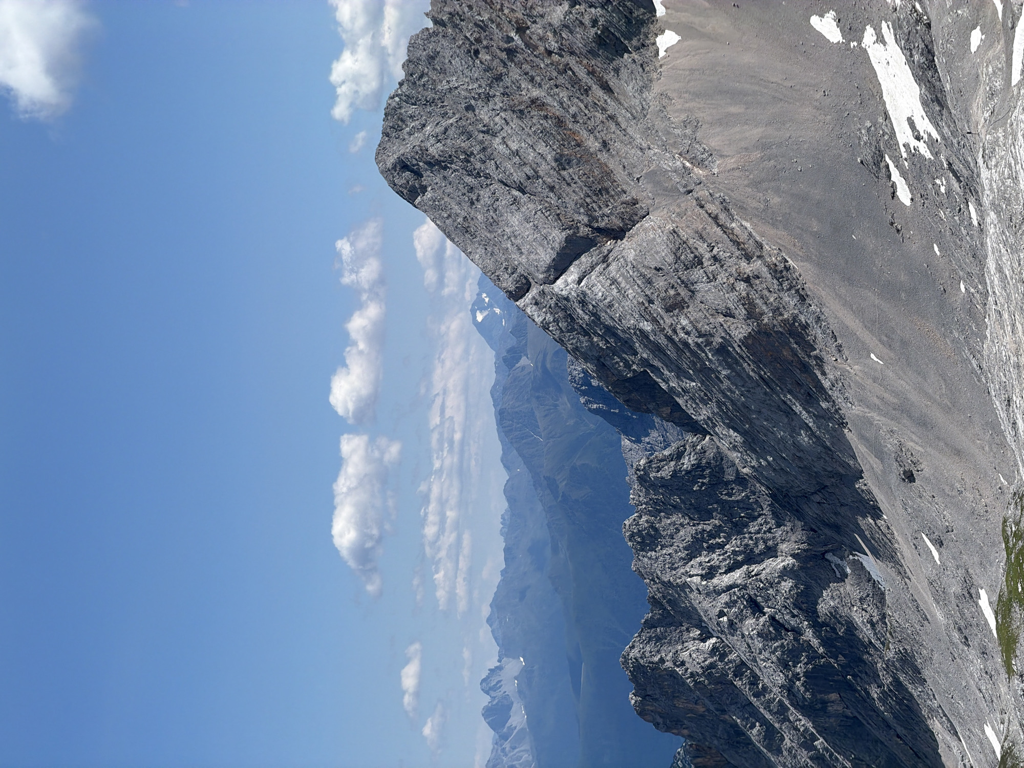 A high-angle, wide shot captures a majestic mountain range under a clear blue sky with scattered white clouds. The foreground is dominated by the rugged, gray, rocky slopes of a mountain, characterized by sharp ridges, steep inclines, and patches of snow. The texture of the rock face is intricate, with visible striations and layers indicating geological formations.
In the mid-ground, a series of layered mountain peaks recede into the distance, gradually fading into a hazy blue. These mountains exhibit softer contours and a more subdued color palette, suggesting a greater distance and atmospheric perspective. Some of these distant peaks also show hints of snow or ice.
The sky occupies the upper portion of the image, a vibrant, cloudless blue with a few wispy white clouds adding depth. The lighting suggests a bright, sunny day, casting distinct shadows and highlighting the topographical features of the mountains. The scene is devoid of any human presence or artificial structures, presenting a raw and untouched natural landscape.
The image is identified as being from the Zugspitze massif, which is part of the Wetterstein mountain range and is located on the border between Germany and Austria, near the Bavarian town of Garmisch-Partenkirchen, Germany. The dominant geological formations are sedimentary rocks, primarily limestone, which contribute to the sharp and angular features of the landscape. The presence of snow patches indicates a high altitude environment, likely above the tree line.