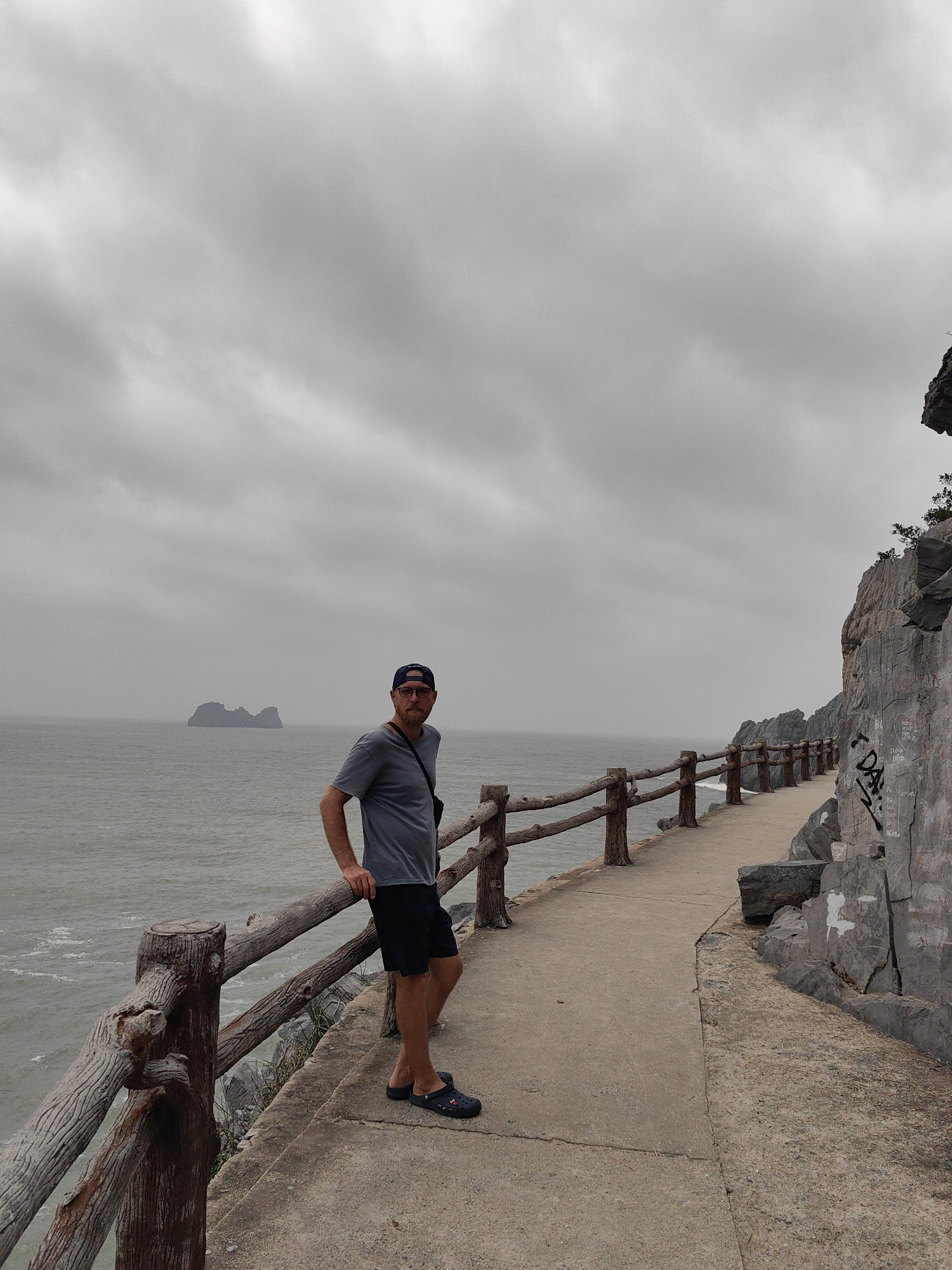 A man stands on a concrete path in Hải Phòng, Vietnam. The path is lined with a wooden railing on the left and a rocky cliff face on the right. The sea stretches out to the left, and a small island is visible in the distance. The sky is overcast. The man is wearing a gray t-shirt, dark shorts, a baseball cap, and Crocs. He is leaning against the railing, looking toward the camera. Graffiti is visible on the cliff face.