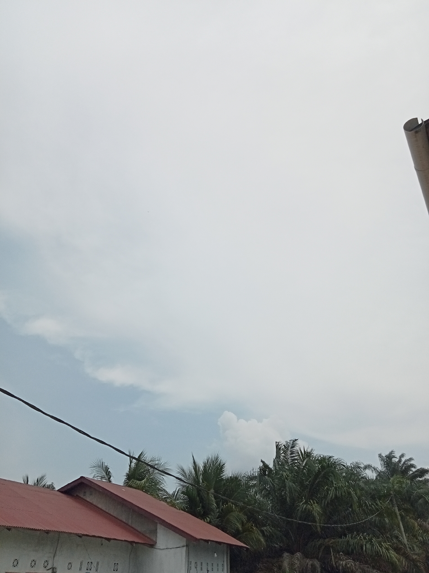 The image shows a view of the sky and rooftops of buildings in Melayu Besar Kota, Indonesia. The sky is overcast with some light clouds visible. In the foreground, there are the red metal roofs of two adjacent buildings. These buildings appear to be single-story structures with white walls and decorative window openings. Trees, likely palm trees, are visible behind the buildings, their dark green fronds contrasting with the lighter sky. A power line runs diagonally across the upper portion of the image, partially obscuring the view of the sky and trees. On the far right side of the image, a portion of a vertical, cylindrical object, possibly a smokestack or part of a tower, is visible. The scene suggests a semi-urban or rural environment, common in many parts of Indonesia.