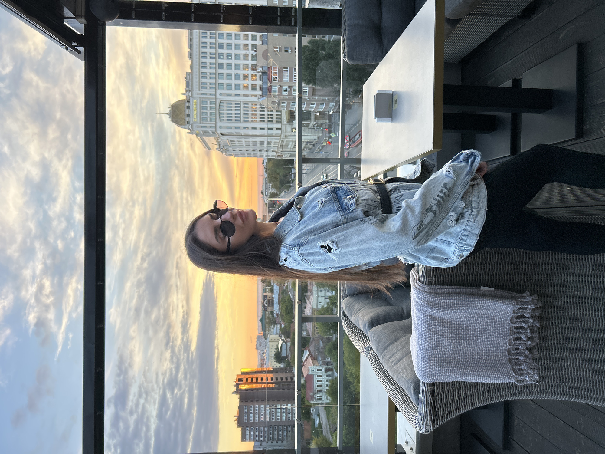 A woman is sitting on a wicker chair on a balcony overlooking a city at sunset. She is wearing sunglasses and a denim jacket. The sky is filled with dramatic clouds and the warm colors of sunset. In the background, there are buildings and a street with cars. The overall mood is serene and relaxed.
The woman has long, dark hair and is wearing a light-wash denim jacket with distressed details. She has on dark sunglasses and appears to be looking towards the sunset. Her posture is relaxed as she leans back in the chair.
The setting is an outdoor balcony or patio with a view of a city. There is a table to her right with a small white object on it, possibly a tissue dispenser or ashtray. A grey blanket is folded on the chair next to her. The architecture in the background includes a large, ornate building with a golden dome, reminiscent of historical European architecture. Other buildings are more modern, with visible windows. The sky shows the vibrant colors of sunset, with streaks of orange, yellow, and pink. The time of day is clearly sunset.
There is some text visible on the golden-domed building. It appears to be in Cyrillic script and reads "КОШЕЛЕВ". This likely refers to the name of the building or a company associated with it. The location is identified as Novokuybyshevsk, Russia.