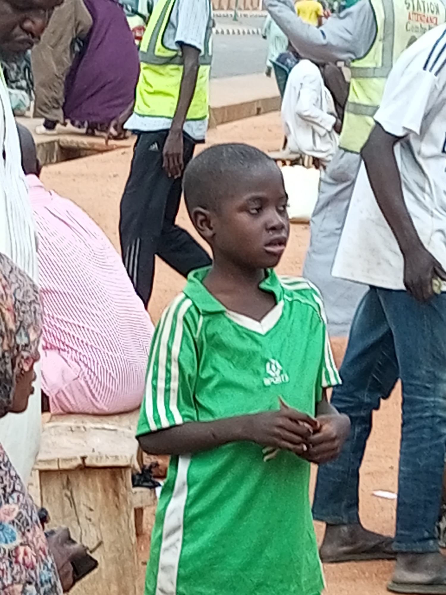 This is a medium shot of a group of people in what appears to be a market or busy street area in Gombe, Nigeria. In the foreground, a young boy is looking to his left, wearing a green t-shirt with white stripes on the sleeves and a white trim around the neck. His hands are clasped in front of him. To his left, a person is wearing a pink and white striped garment. Behind the boy, a person wearing a bright yellow vest over a white shirt and black pants is visible. Further in the background, other individuals are present, some wearing traditional Nigerian attire, and a building with signage indicating "STATION" is visible. The ground is reddish-brown earth, suggesting an outdoor environment.