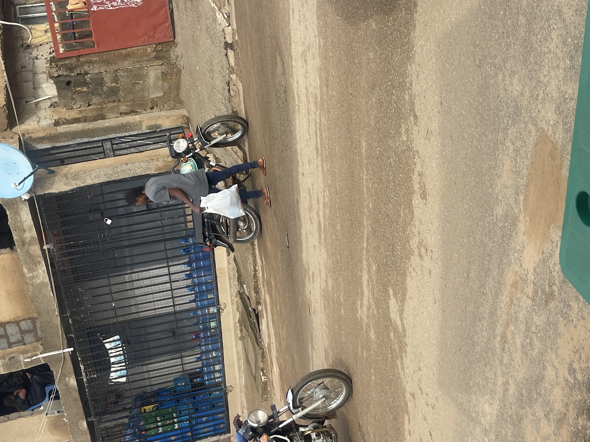 A person is walking on a paved street in front of a building with barred windows. The person is carrying a white plastic bag and is wearing a grey t-shirt and blue jeans. A motorcycle is parked next to the person. The building appears to be a shop or residence, with stacks of blue plastic water bottles visible behind the bars. The street is worn and cracked in some areas. The overall scene suggests an urban environment, possibly in Abuja, Nigeria, given the water bottle branding if any were discernible.