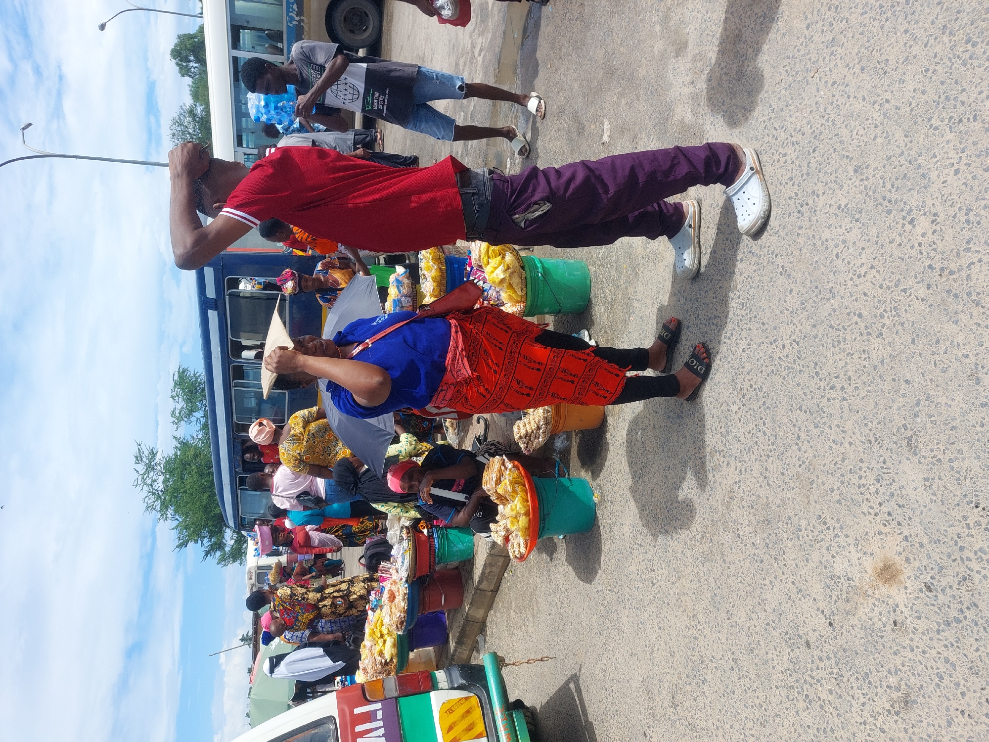 This image captures a vibrant, bustling street scene in Dar es-Salaam, Tanzania, under bright daylight with a partly cloudy sky. The perspective is slightly elevated, looking down into the street.
The scene is densely populated with people and activities, suggesting a market area or a busy public transport hub. In the foreground, a man wearing a red polo shirt, purple trousers, and white Crocs stands with one arm raised to his head. Beside him, a woman in a blue top and a red, patterned skirt stands next to several green and red buckets overflowing with packaged snacks, resembling popcorn or puffed rice. She is wearing black leggings and black slide sandals with "DIOR" visible on one.
Further back, numerous other street vendors are visible, some seated, also surrounded by buckets of similar packaged goods. A man in a black t-shirt with "LIVING THAT" and "NEW YORK" visible text, and denim shorts, is seen holding a large blue multi-gallon water bottle.
Several buses are parked or moving in the background, indicating a transport-oriented setting. People are congregated around these buses. At the very bottom edge of the frame, the front of a multi-colored vehicle, possibly a tuk-tuk or Bajaj, is partially visible, with "TRAVEL" or similar text on its side. Street lamps extend into the sky. The ground is paved asphalt. The overall atmosphere is one of lively commerce and daily urban activity.