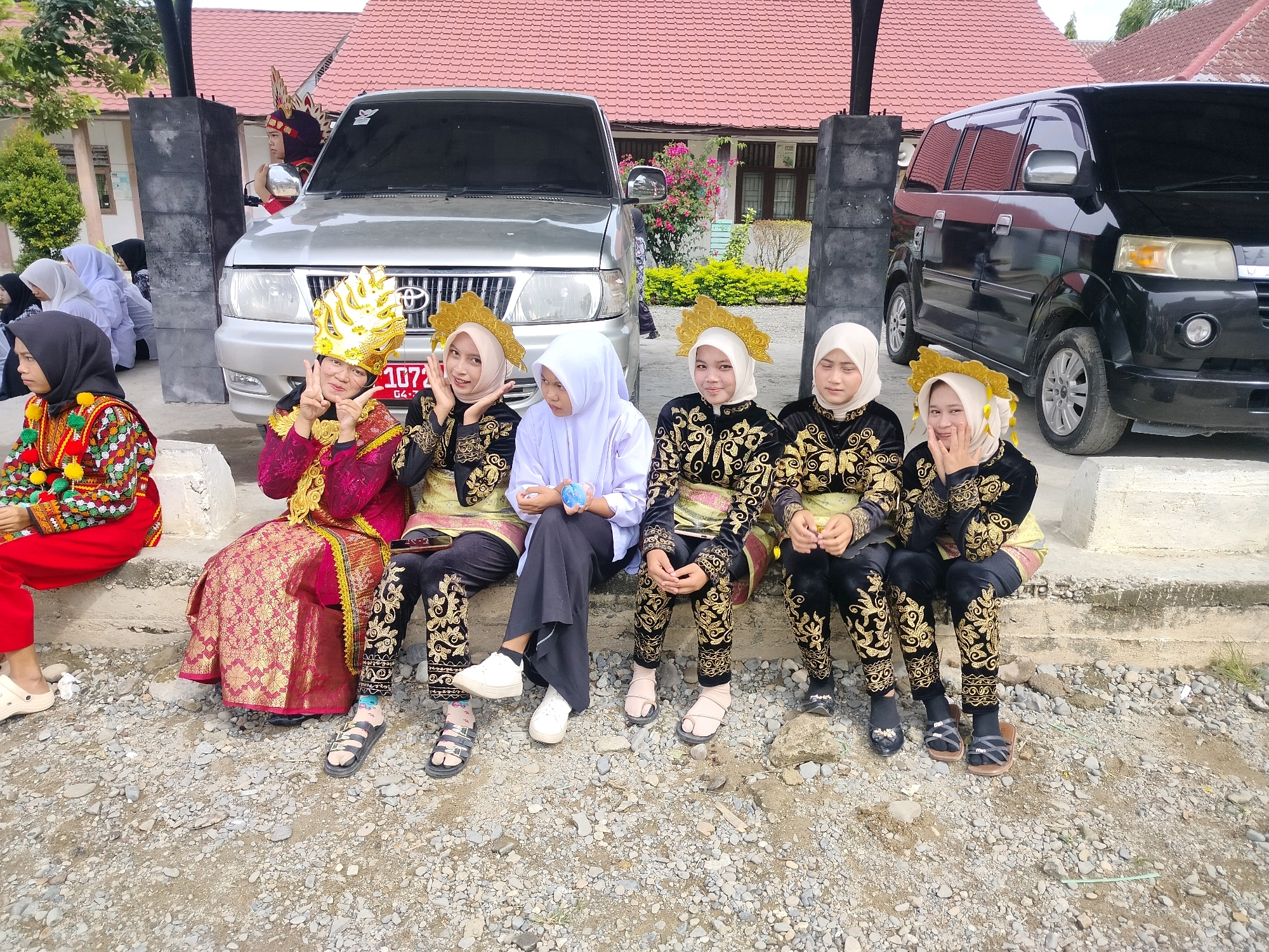 In Purwodadi, Indonesia, a group of six young females is seated on a concrete ledge outdoors, posing for a photograph. Two silver cars are parked behind them. The females are dressed in traditional attire, with three wearing ornate gold headdresses and black outfits adorned with gold embroidery, while another wears a white hijab and matching black pants, and another is dressed in a vibrant red outfit with embroidered details. The group appears to be participating in a cultural event or performance. The environment consists of a paved area with loose gravel, bordered by a building with a red-tiled roof in the background.