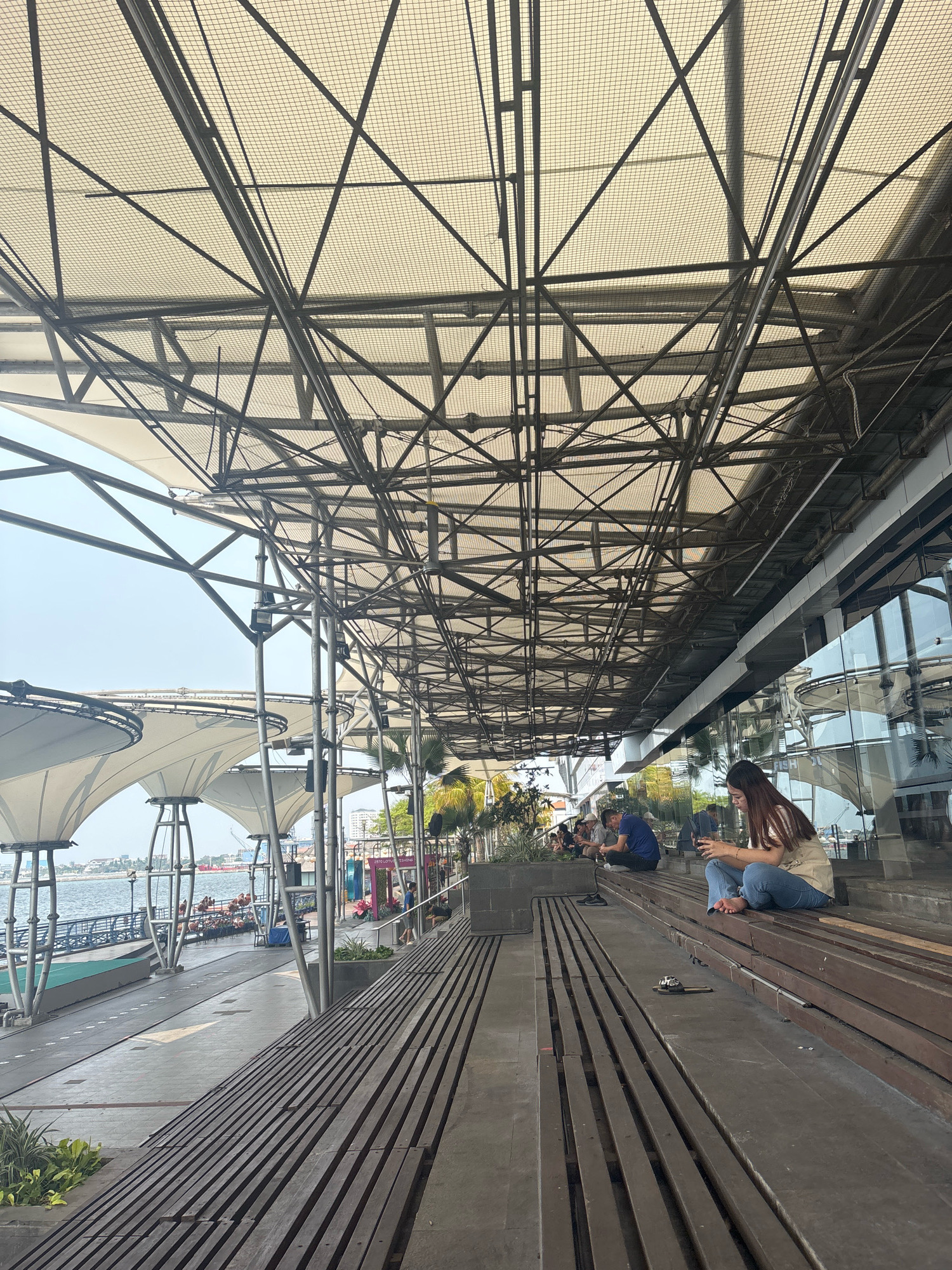 The image depicts a waterfront area in Ancol, North Jakarta, Indonesia. A large, modern canopy with a complex metal framework and a mesh ceiling covers a wide seating area constructed from wooden planks. In the foreground, several people are seated on the wooden steps, some looking at their phones. To the left, a promenade runs alongside the water, with more seating and white tent-like structures. The water appears calm, with a few boats visible in the distance. The overall atmosphere is relaxed and public, suggesting a commercial or leisure hub.