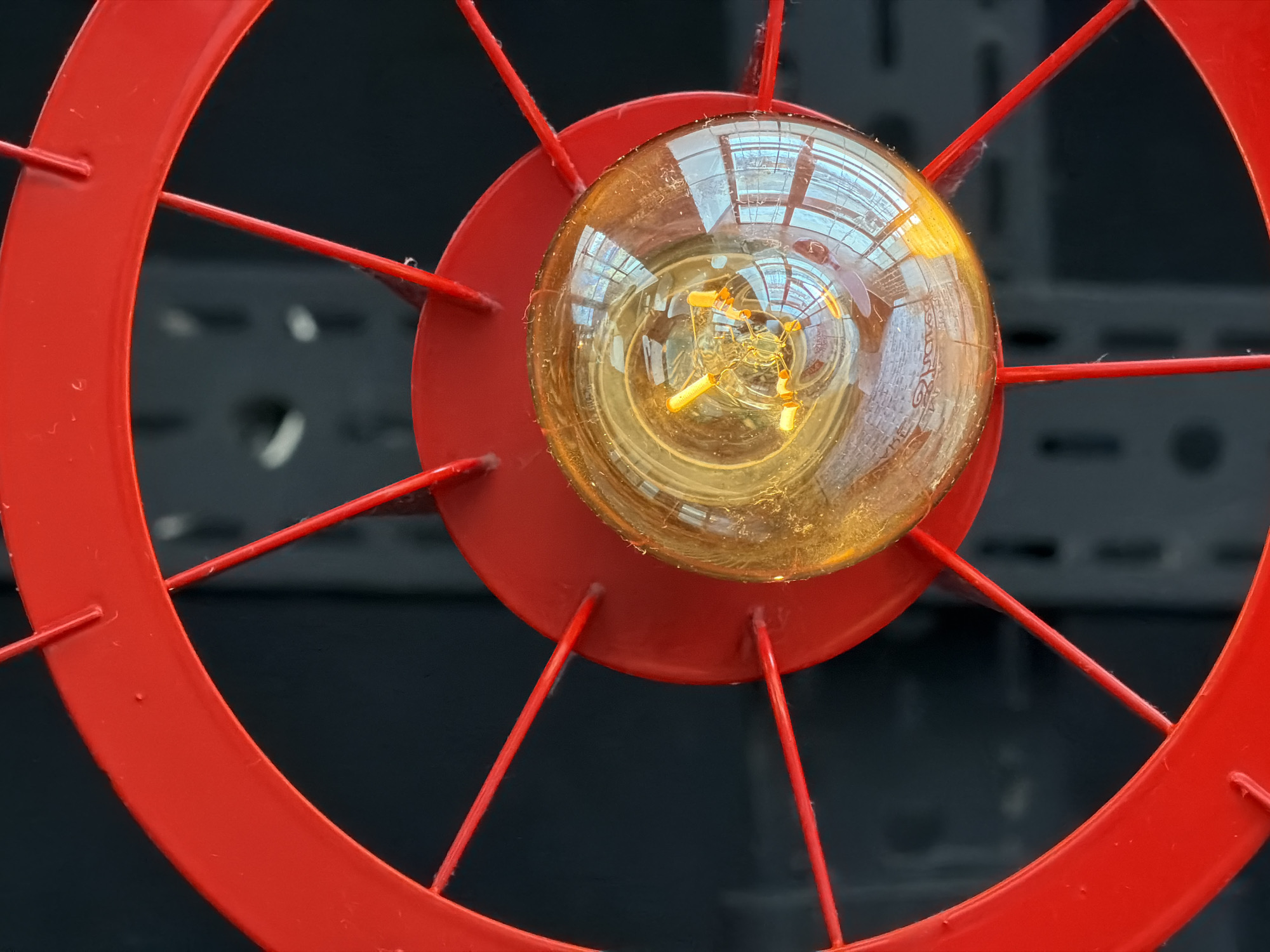The scene captures an illuminated, amber-tinted, vintage-style incandescent light bulb mounted within a bright red, spoked metal fixture, resembling a wheel or fan guard, situated in Chaikovsky, Russia. The fixture features a central hub, slender spokes, and a prominent outer rim. The spherical glass surface of the bulb reflects interior filaments and external environmental elements, including what appear to be window frames or structural components rendered in light blue and white hues. Minor dust accumulation is visible on the bulb's surface and the red fixture. The background is dark, out-of-focus, and reveals indistinct vertical elements consistent with a metal shelving unit or perforated panel. No individuals or active interactions are present. The perspective is upward-looking, focused on the light source.