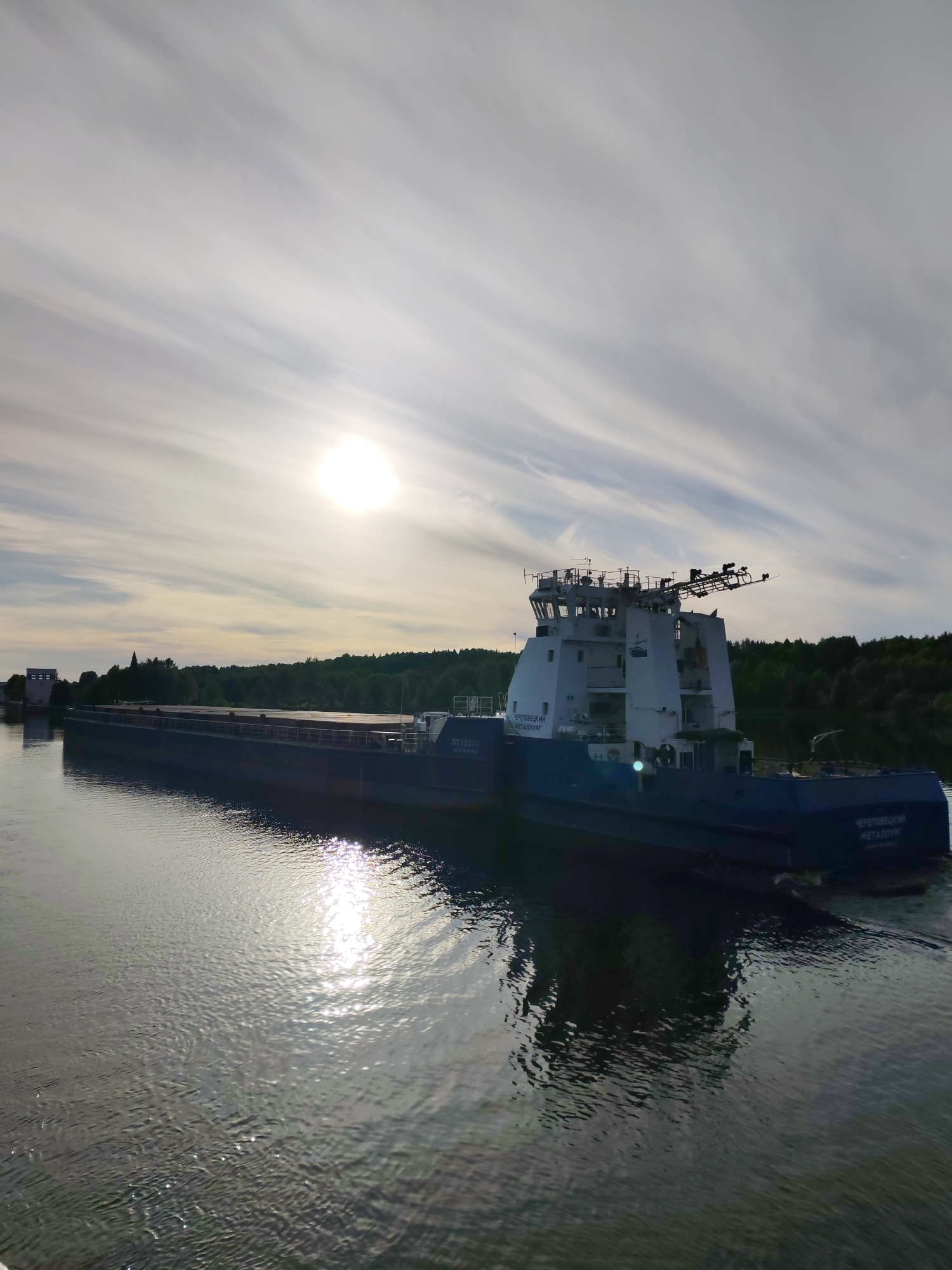 A large, dark blue and white cargo vessel, likely a self-propelled motorship or a barge with a tug component, navigates a tranquil waterway under a bright, partly cloudy sky. The vessel is prominently featured, moving from left to right in the frame. Its white superstructure stands out against the darker hull, with communication or radar equipment mounted on top.
The sun, a brilliant disc, is high in the sky, slightly to the left of center, casting a strong glare and an elongated, sparkling reflection on the water's surface. The sky is streaked with wispy, elongated clouds that diffuse the sunlight.
On the left bank, a light-colored, multi-story building, possibly part of a lock or navigation control station, is visible. Both banks of the waterway are lined with dense, green forests, suggesting an inland river or canal system. The water's surface shows gentle ripples and subtle reflections of the sky and the surrounding environment.
Visible text on the side of the white superstructure and the aft part of the dark blue hull clearly reads "ЧЕРЕПОВЕЦКИЙ МЕТАЛЛУРГ" (Cherepovetsky Metallurg). Additionally, the registration or identification number "ПТ 82018" is visible on the hull below the superstructure. The scene depicts a serene daytime passage in a Russian waterway, with the sun providing dramatic lighting and reflections, likely near Markovo, Russia.