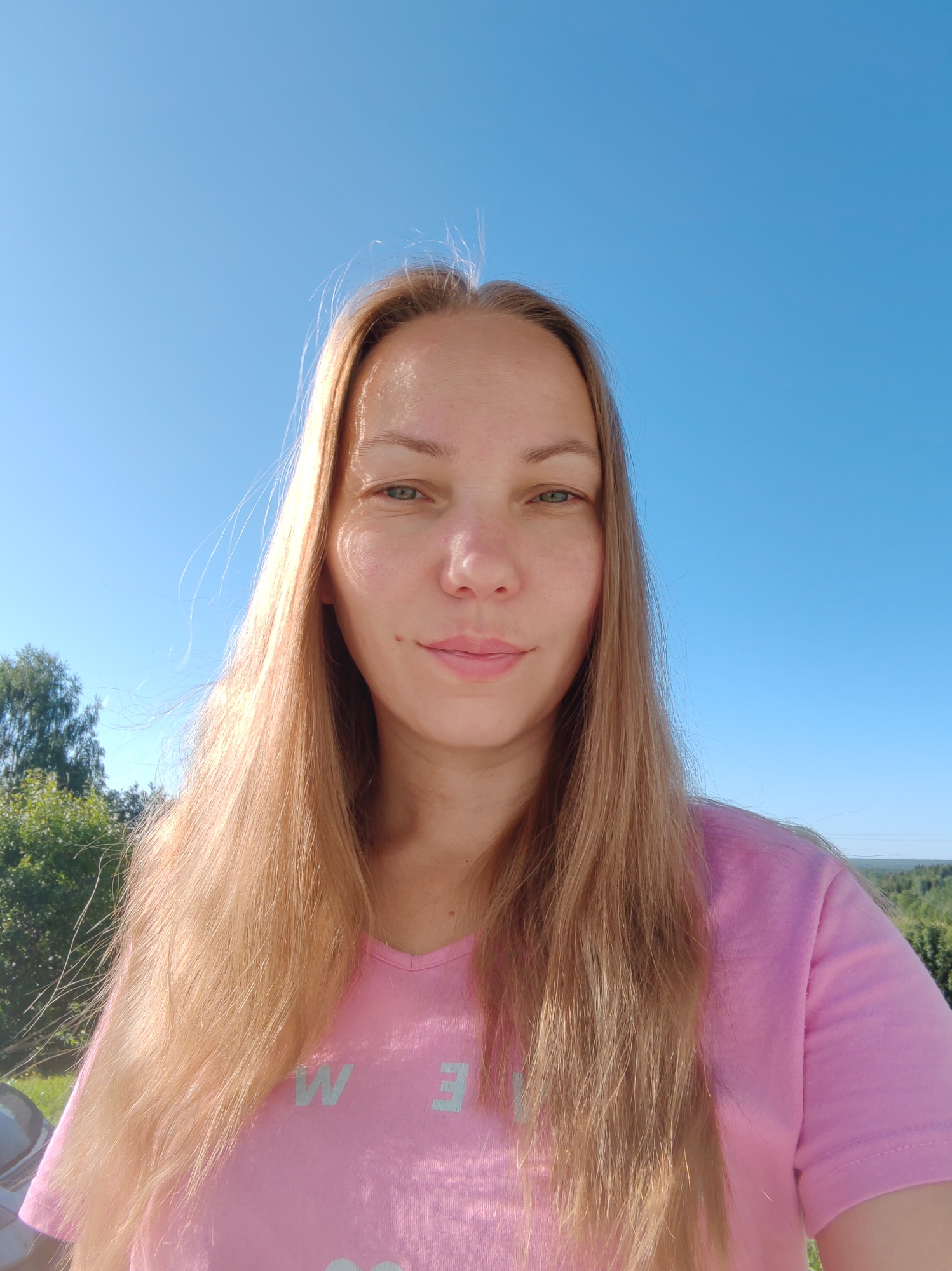 A young woman with long blonde hair and fair skin is pictured in a selfie taken outdoors. She is wearing a pink t-shirt with light blue lettering, partially visible. The background consists of a clear blue sky and a glimpse of green trees and landscape, suggesting a rural or suburban setting. The lighting indicates it is daytime. The location is specified as Chukhlem, Russia.