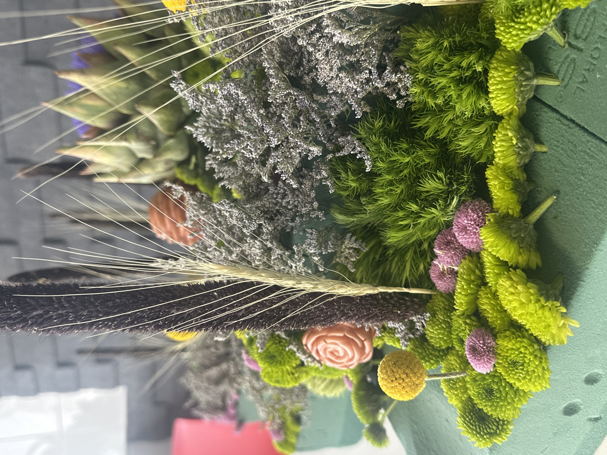 A close-up, vertically oriented shot displays an arrangement of dried and fresh flowers. An artichoke and delicate, wispy seed heads add height. Grayish dried flowers are intertwined with vibrant green, button-like chrysanthemums. A few pink pom-pom flowers and a yellow ball-shaped flower add pops of color. A peach-colored faux rose is present. The flowers are arranged in green floral foam. The context of location in Mexico City is unsupported.