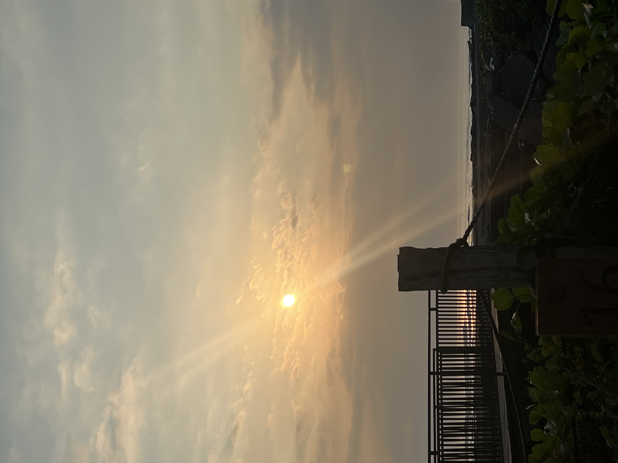 The image depicts a serene beach scene at sunrise, possibly in the area of Mengwi, Indonesia. The sun, a bright orb, is partially obscured by soft, scattered clouds, casting warm rays across the sky. The sea is visible in the distance, with gentle waves lapping the shore. In the foreground, a wooden boardwalk with a dark metal railing leads towards the beach. A weathered wooden post stands prominently in the foreground, with a rope wrapped around it. Lush green foliage is visible at the bottom of the frame, with a partially obscured number "16" on a dark object. The overall atmosphere is tranquil and peaceful, bathed in the soft light of dawn.