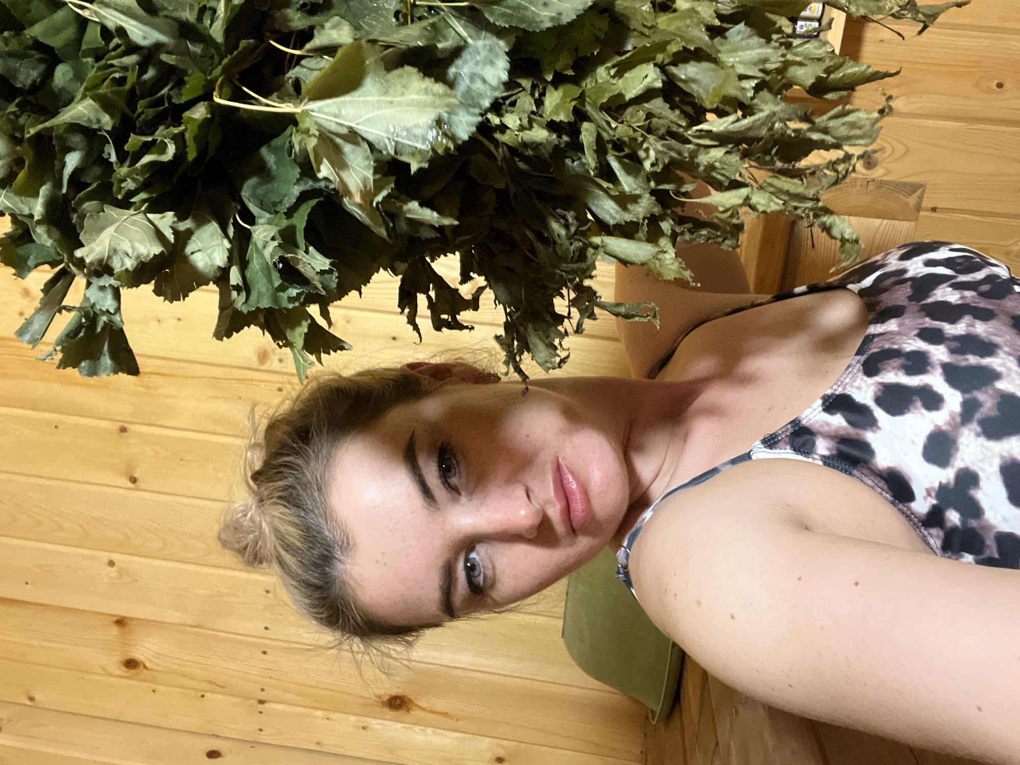 A woman is indoors, lying in front of a wood paneled wall. She has light skin, blonde hair pulled back in a bun, and is wearing a swimsuit with a black and white animal print pattern. A bundle of dried leaves is positioned above her head. The location is Гаревая, Russia.