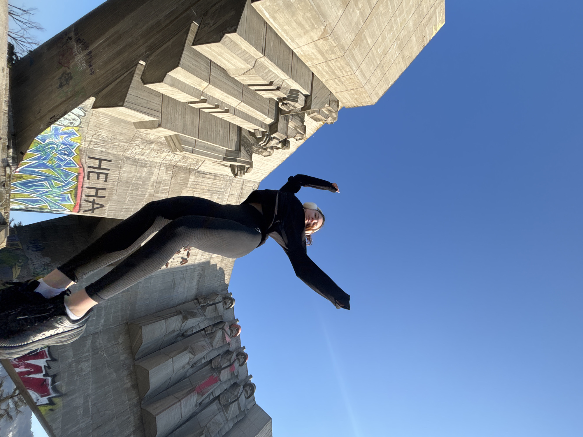 The image is taken from a low angle, pointing upwards towards a monument. A woman is the central subject, positioned in the foreground. She is wearing black athletic clothing with a white headband and is leaning backwards, appearing to be performing a gymnastic pose or stretch on the monument. The monument itself is large, made of grey concrete, and covered in graffiti in some areas. The sky is clear and blue and serves as the backdrop. The location is the Monument of the Bulgarian-Soviet Friendship in Varna, Bulgaria.