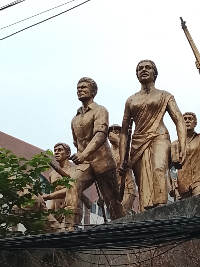 The image displays a large, multi-figure bronze-colored sculpture, identified as the "Shadhinota Shongram" (Freedom Struggle) monument, located on the campus of the University of Dhaka, Dhaka, Bangladesh. The sculpture depicts a dynamic group of individuals, predominantly men and women, cast in a matte, aged bronze-like finish, striding forward with determined expressions.
In the foreground, slightly left of center, a male figure in a collared shirt and trousers is depicted in mid-stride, looking upwards and to his left. To his immediate right, a female figure dressed in a sari holds a long rifle-like object vertically in her right hand and a rectangular object, possibly a book, in her left, also looking forward with a resolute gaze. Behind and around these central figures are additional statues, some carrying similar weapon-like objects, contributing to the impression of a unified march or struggle.
The base of the sculpture, made of concrete or stone, is partially visible. Multiple black electrical or communication wires are strung horizontally across the lower mid-ground, slightly obscuring the bottom portion of the monument. A cluster of green foliage from a tree or bush is visible on the left side, partially in front of some of the figures. In the background, a brown-tiled building with a sloping roof is discernible on the upper left, while the majority of the upper frame consists of a light, overcast sky. A vertical pole-like structure extends upwards from the top right edge of the frame. The perspective is a low-angle shot, looking up at the monument.