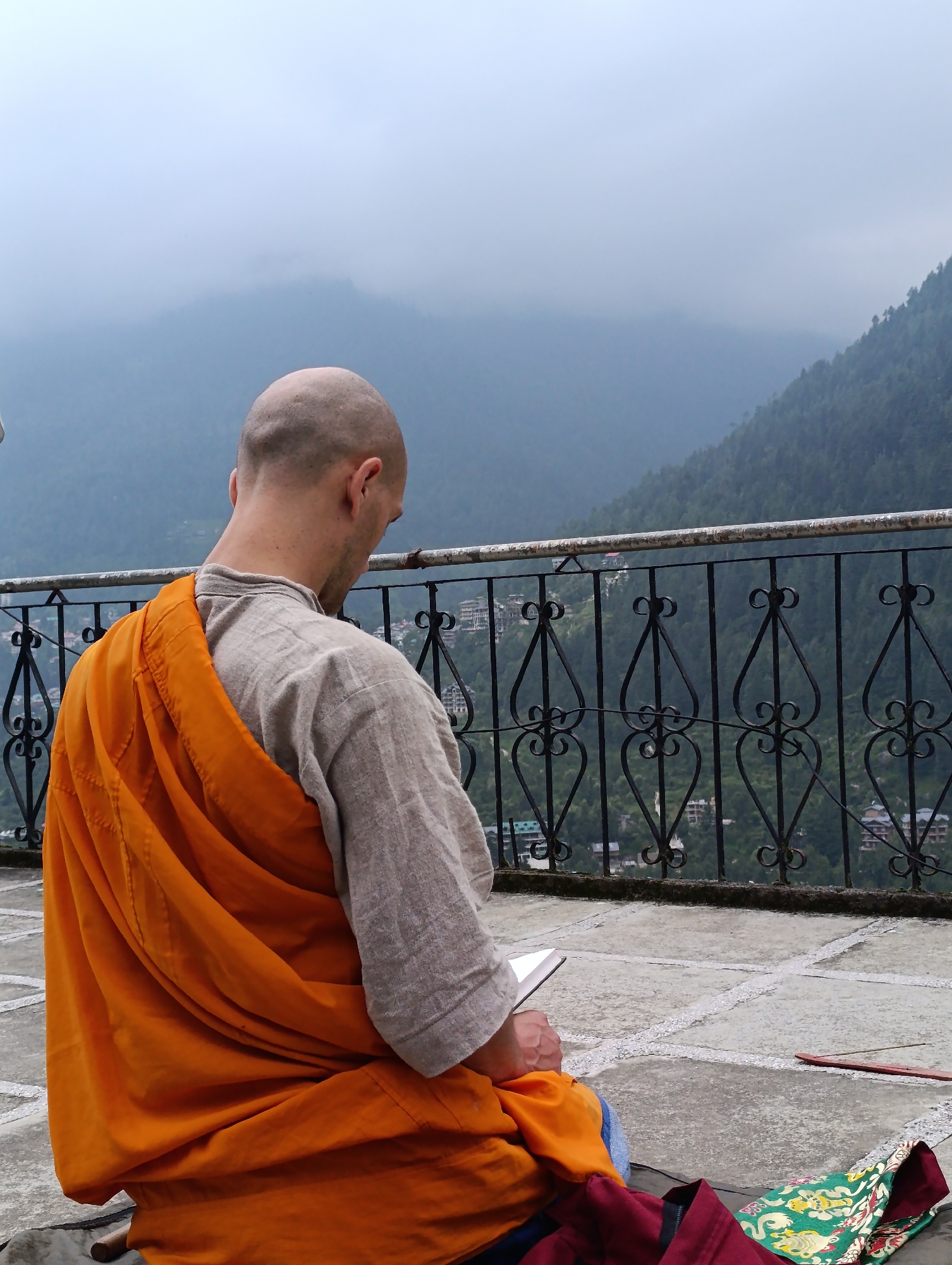 A person, likely a monk, sits facing away from the camera on a stone patio. They are wearing a simple, light-colored tunic and an orange robe draped over their shoulders. The monk is holding a book or tablet and appears to be reading or studying.
In the background, a black wrought-iron railing with decorative scrollwork separates the patio from a steep, densely forested mountainside. Below the railing and on the mountain slopes, small buildings and structures are visible, suggesting a village or town nestled in the valley. The overall atmosphere is serene and contemplative.
The weather appears overcast and misty, with the mountain peaks shrouded in fog, creating a soft, diffused light. This suggests it might be morning or late afternoon, or simply a cloudy day. The setting, with its mountainous landscape and the monk's attire, points to a location in the Himalayas, consistent with Vashisht, India. The presence of a colorful embroidered cloth and an incense stick on the patio suggest ongoing spiritual or ritualistic activity. No visible text is present in the image.