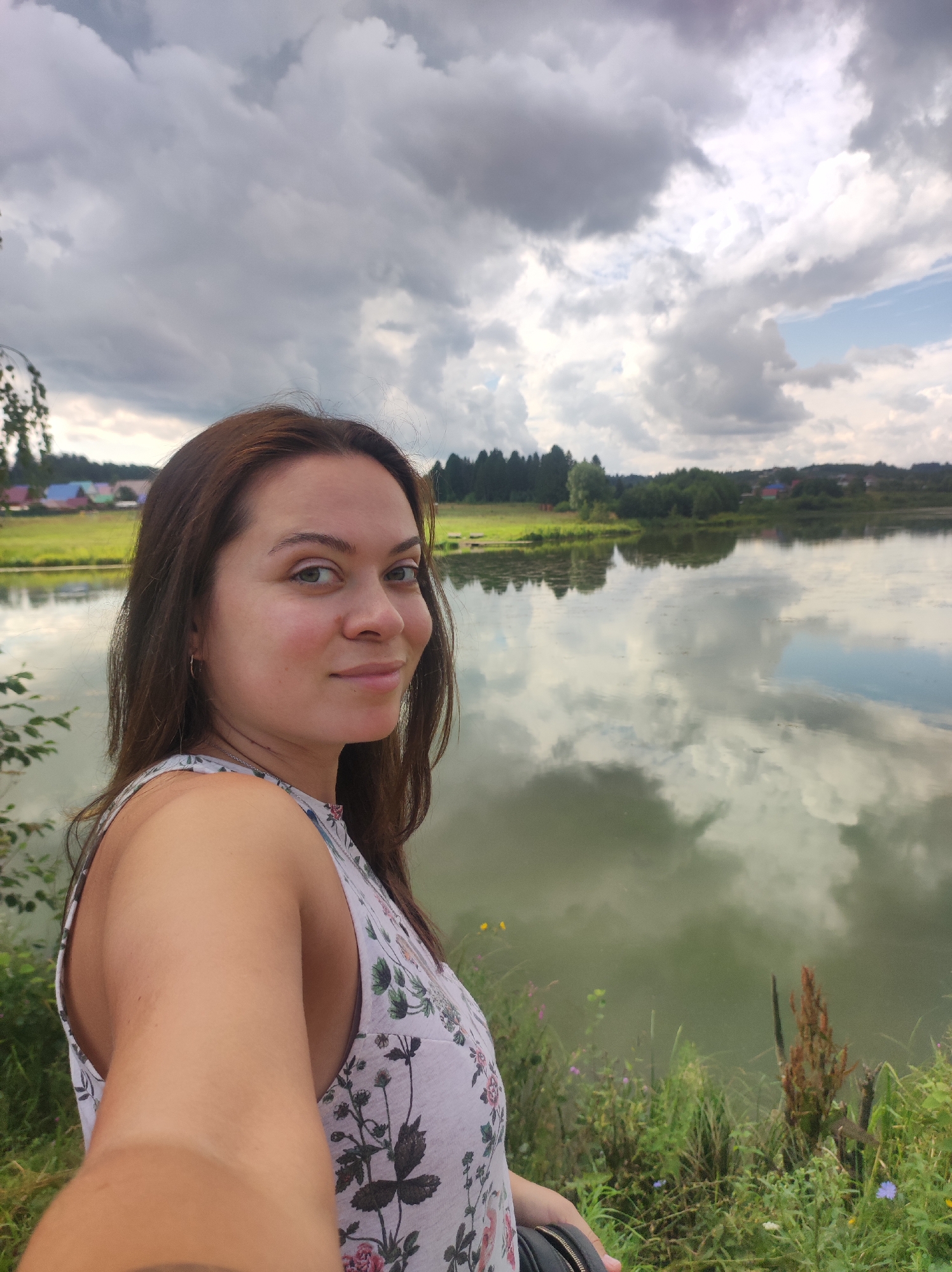 A woman with brown hair is captured in a selfie, her head and upper body taking up the left side of the frame. She is wearing a white dress with a floral print. Her gaze is directed slightly away from the camera, and she has a subtle smile. Behind her, a tranquil body of water reflects the dramatic, cloudy sky. The background features a lush green landscape with trees and distant colorful buildings, suggesting a rural or semi-rural setting, possibly on the outskirts of Фоки, Russia. The overall atmosphere is serene and natural.