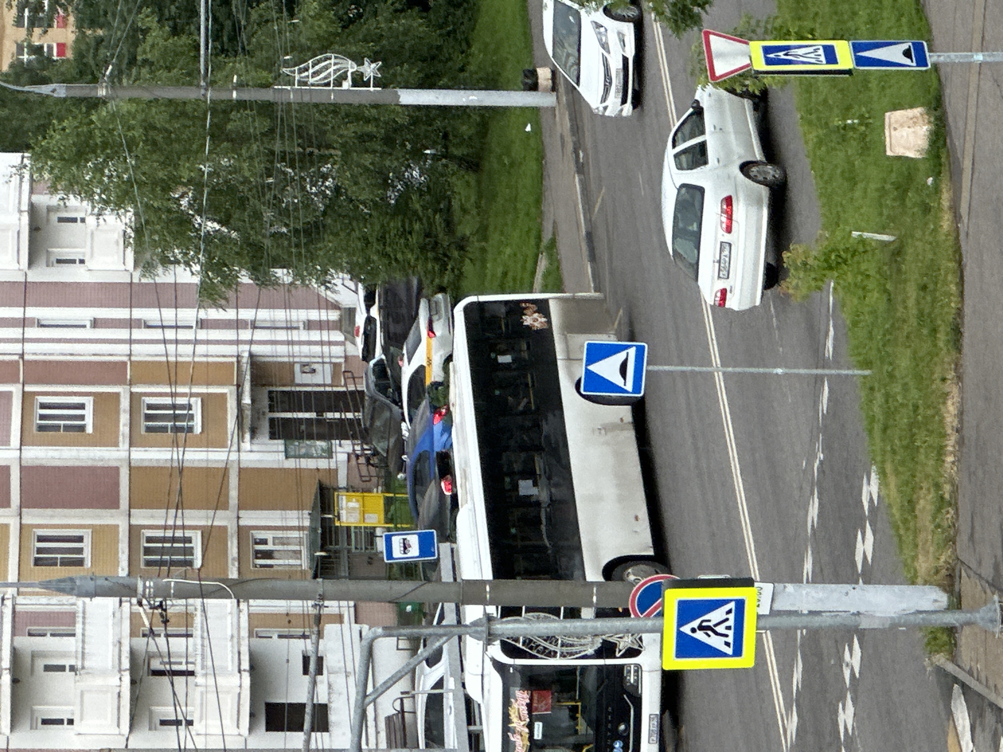A wide-angle, eye-level outdoor shot captures a street scene in Podolsk, Russia. The foreground is dominated by a silver sedan driving away from the viewer, with a pedestrian crossing signpost to its left. The signpost features a yellow square with a pedestrian symbol and a blue square with a white upward-pointing triangle. Behind the sedan, a white bus is visible, its front on the left side of the frame. The bus has writing on its windshield and side. Further back, a blue car is parked. The background includes a multi-story building with a facade of beige and pink sections, several windows, and balconies. Trees with green foliage are present to the right of the building. The street is paved, with white lane markings visible. The sky is overcast.
