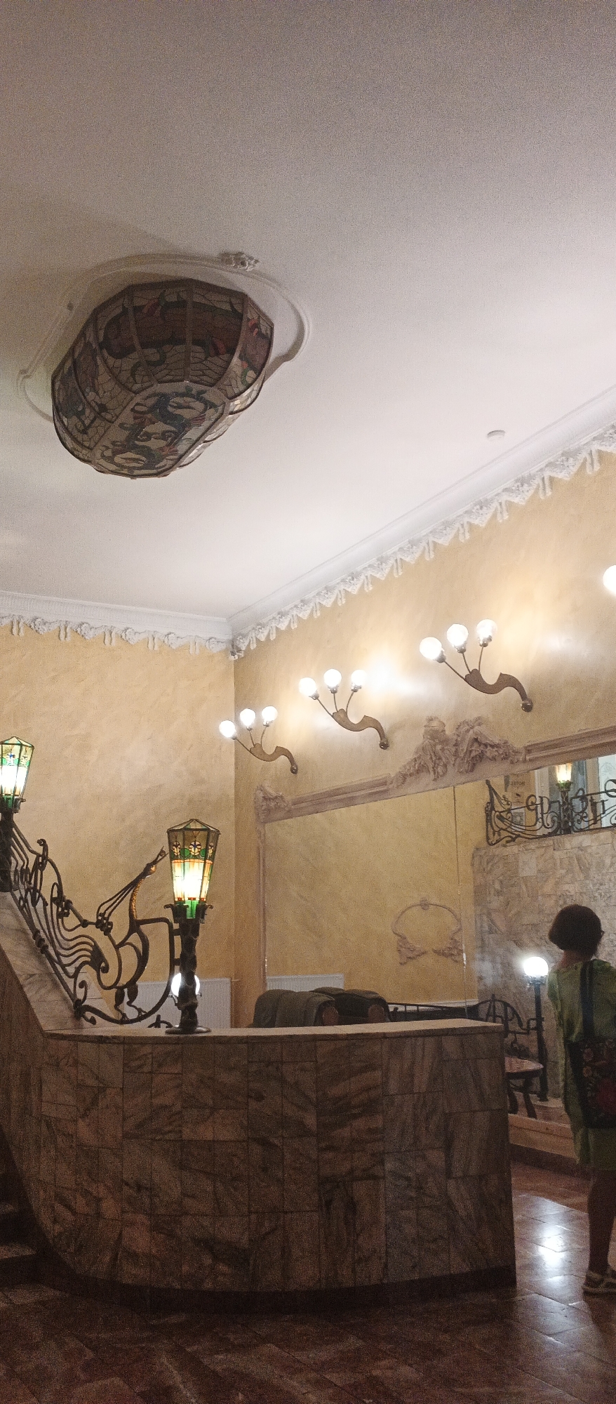 The image depicts the interior of a building, likely a hotel or a public establishment, featuring an ornate staircase with stained-glass lamps, decorative wall sconces, and a large mirror. The walls are a pale yellow, and the ceiling features intricate moldings. A person in a green shirt is visible in the lower right of the frame, facing away from the camera. The staircase has a marble base and elaborate metal railings. The overall impression is one of historical elegance. This location is identified as the guest house of the Palanok Castle in Berehove, Ukraine.