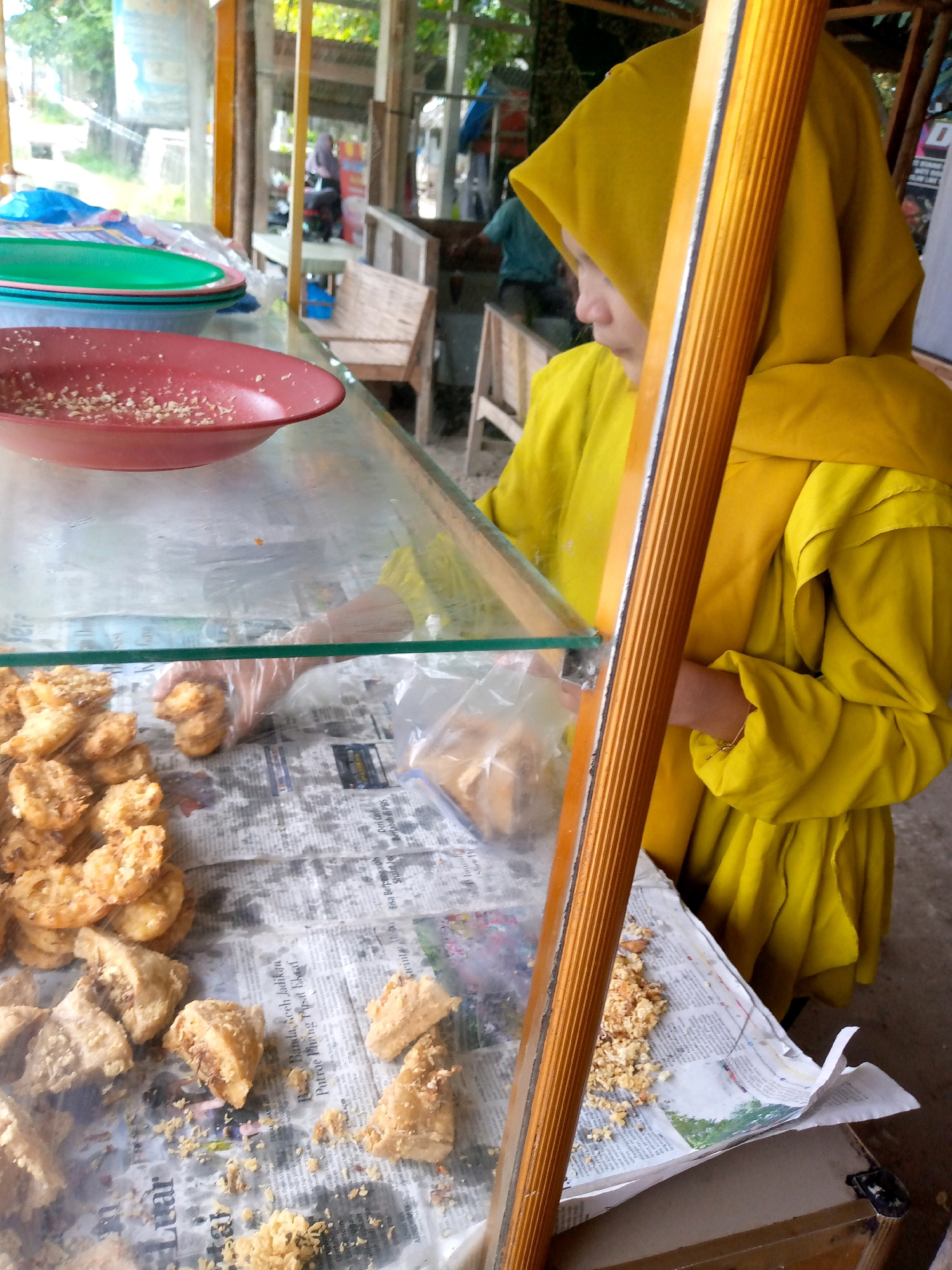 An outdoor food stall in Keudebayu, Indonesia displays fried snacks. A woman wearing a yellow hijab and dress stands behind the stall, holding a fried snack in a clear plastic glove. Several piles of fried snacks, some triangular and some circular, are arranged on a newspaper-covered surface inside a glass display case. A red bowl with food remnants sits on a shelf above the snacks, with green and blue plastic plates stacked behind it. Wooden benches and a building with a corrugated metal roof are visible in the background.