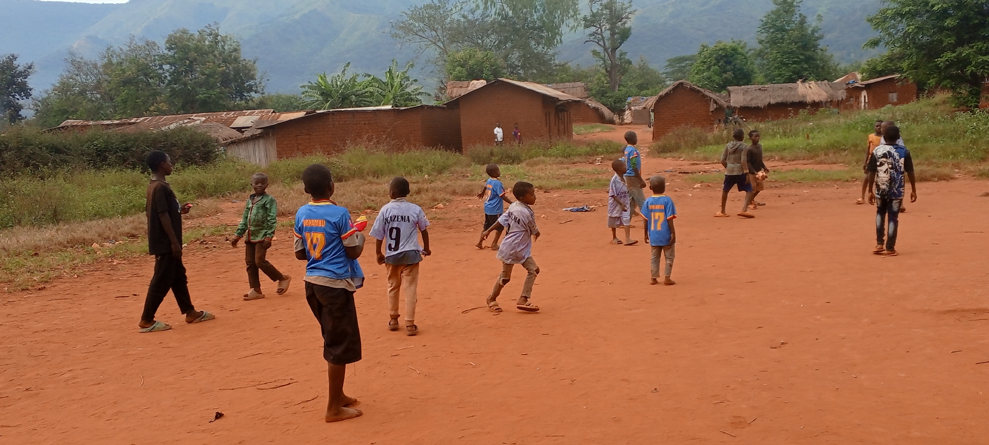 The image shows a group of children playing on a dirt field in Ngalula, Democratic Republic of the Congo. Several small brick and mud houses with metal roofs are visible in the background, along with trees and a distant mountain range under a cloudy sky. The ground is a reddish-brown color. The children are of African descent and appear to be engaging in a game, possibly soccer or a similar sport. Some are wearing jerseys with names and numbers. The scene conveys a sense of everyday life in a rural setting.