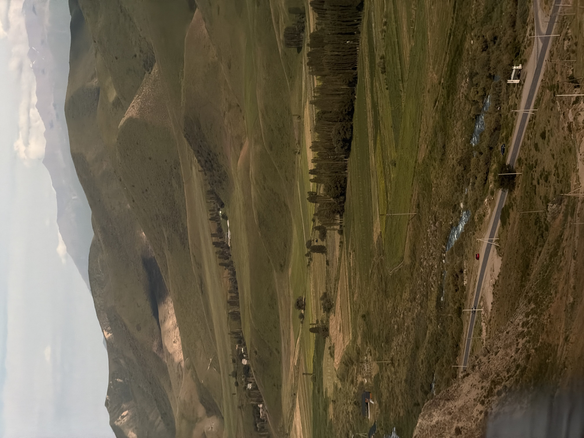 This is an aerial shot of a mountainous, rural landscape in Karagai Bulak, Kyrgyzstan. The scene is dominated by rolling green hills and valleys, with a winding road cutting through the terrain. A small, blue-roofed building is visible near the road, along with a red car parked on its side. Further down the road, a white structure resembling a house with a brown roof is also present. A narrow, rocky stream flows alongside the road in some sections. The hills are covered in sparse vegetation, with a line of trees marking a distinct boundary on one of the slopes. The sky is overcast, suggesting a cool, possibly windy day, with diffused light. The overall impression is one of a remote and natural setting, likely captured during daylight hours. There is no visible text in the image.