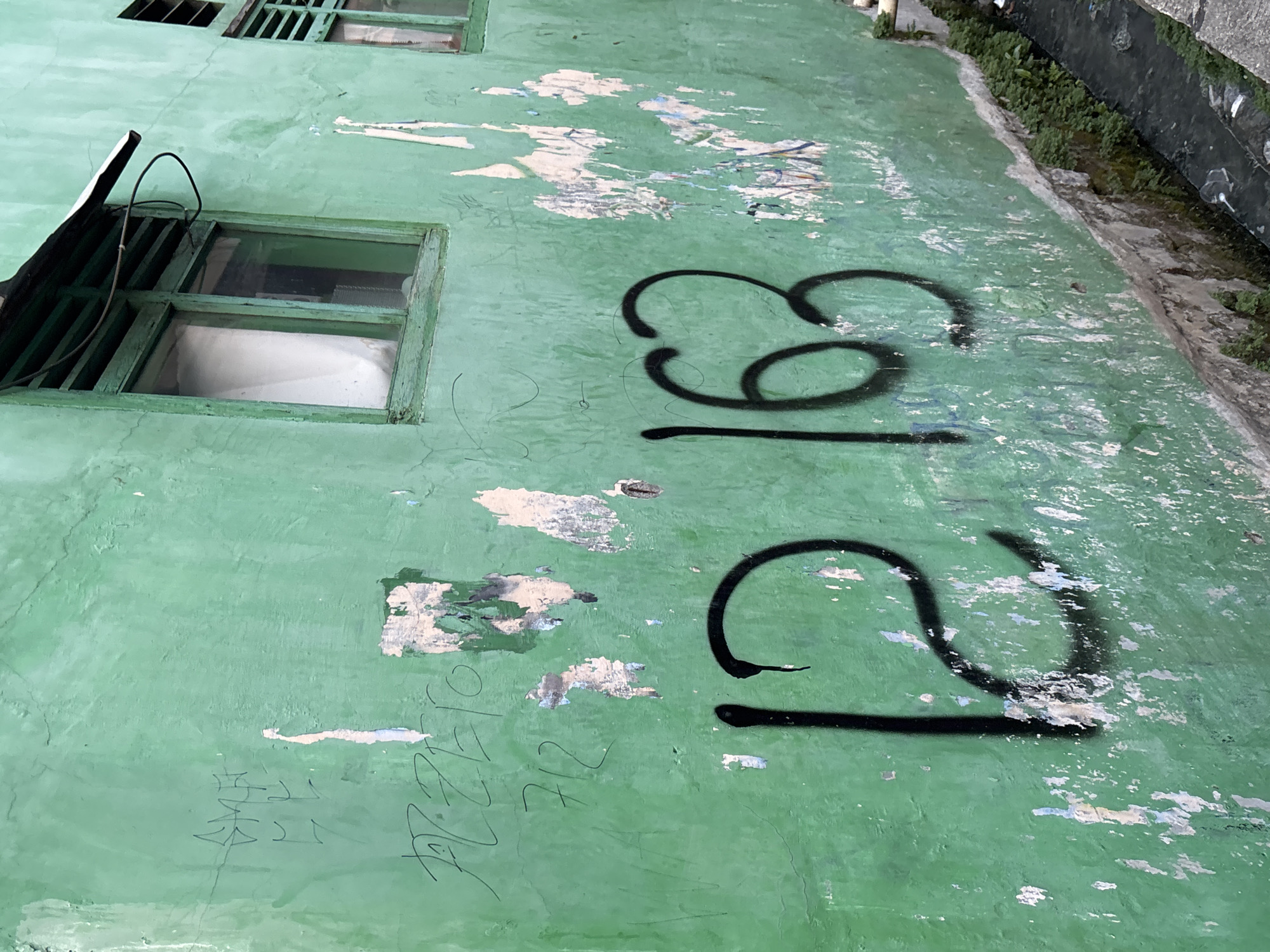 The image depicts a close-up, eye-level exterior shot of a weathered, bright green wall in a gritty urban environment. This location is identified as being in North Jakarta, Indonesia. The wall exhibits significant wear and tear, with patches of peeling paint and underlying plaster visible. Graffiti is present on the wall in several areas. Most prominently, large black spray-painted numbers "12" and "163" are visible. Above these numbers, other faint markings and what appear to be handwritten tags or names are discernible, rendered in a darker, possibly faded paint. The wall has two windows with green wooden frames, one larger and one smaller. The larger window has a white curtain visible inside. A thin metal awning extends from the top of the larger window, and a black wire is loosely draped over it. The lower portion of the wall meets a concrete ledge, below which a narrow, dark, and somewhat dirty drainage channel runs parallel to the wall, lined with small green plants. Debris, including plastic, is present in the drainage channel. The overall impression is one of a neglected urban facade.