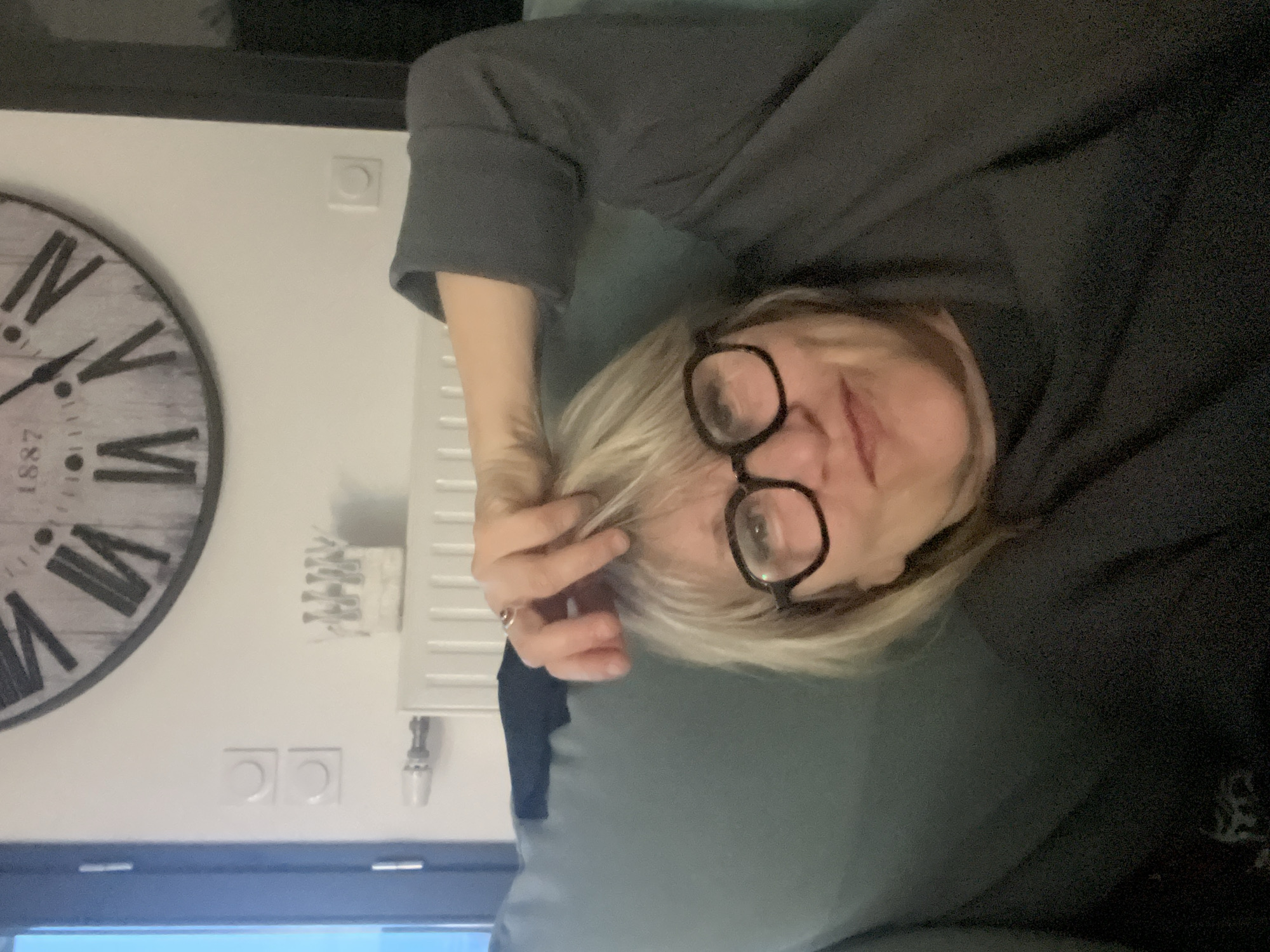 The image depicts a woman with blonde hair and glasses lying on a gray or green surface. She is wearing a dark gray top. Her right hand is near her face, appearing to touch her hair. In the background, there is a wall with a large, circular clock displaying Roman numerals and the year 1887. Also visible are a white radiator, electrical outlets, and a small decorative item on the wall. The location, Chambœuf, France, is not verifiable from the image content alone.