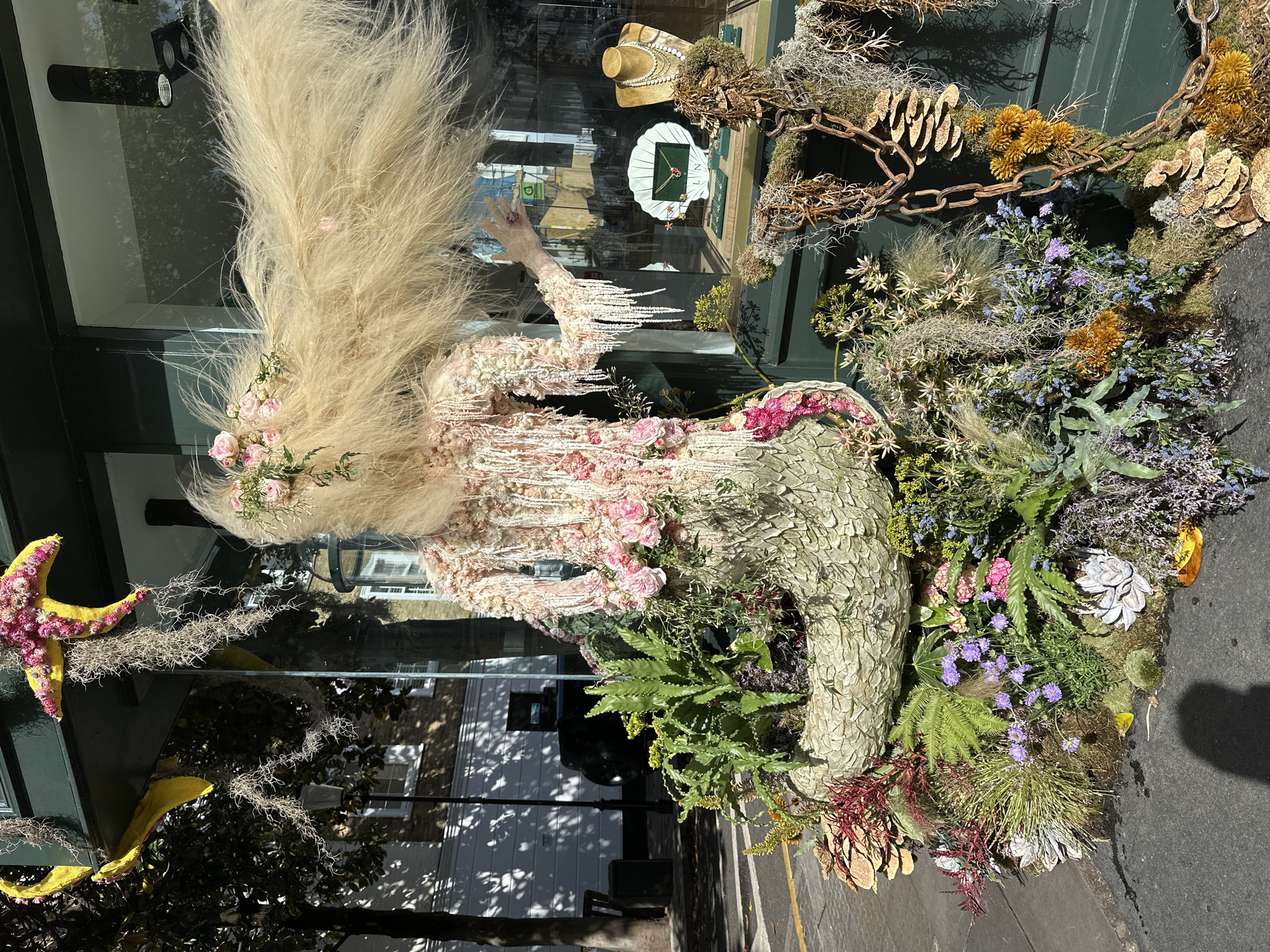 A window display in London, United Kingdom shows an elaborate floral arrangement and mannequin inside of a glass window. The mannequin is adorned with feathers and pink floral embellishments, with its arm raised. Adjacent to the mannequin is a large, round floral arrangement featuring ferns, moss, and various purple, pink, and green flowers. A chain is placed atop the arrangement. A yellow starfish is attached to the upper left corner of the window. Reflected in the window are building facades and trees. Inside the window, a display of jewelry is visible, with a mannequin wearing a pearl necklace and a white oyster shell with a green necklace.
