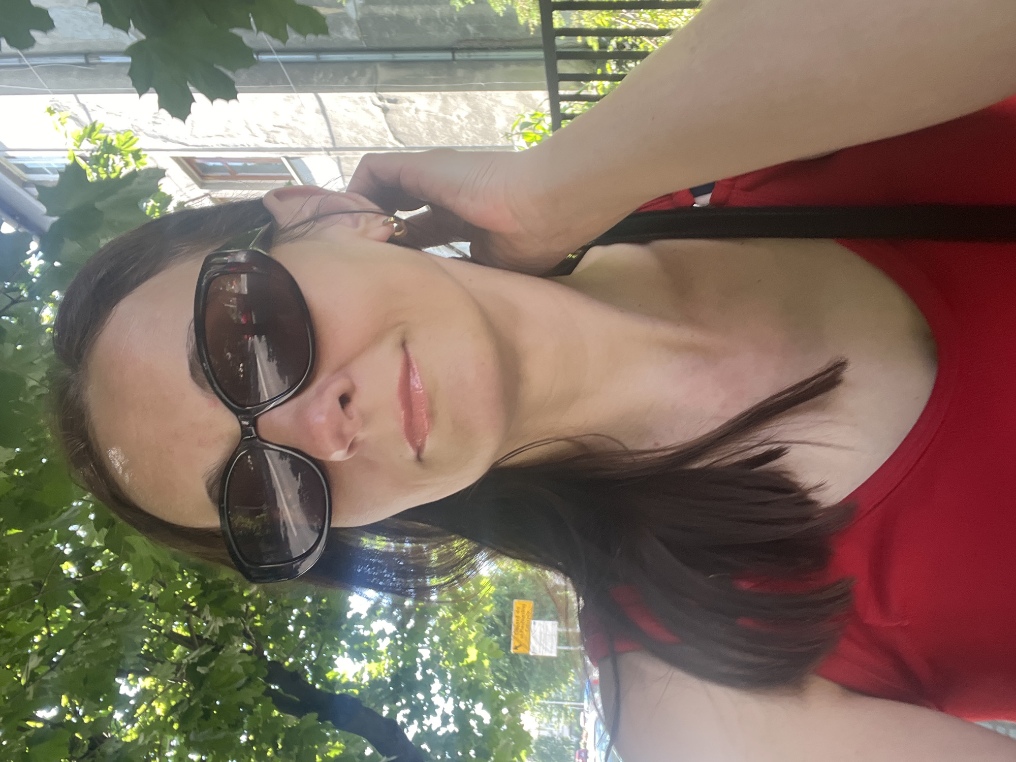 A woman in sunglasses and a red top stands outside, with trees and a building in the background. The woman is facing the camera and has her right hand raised to her ear. She has medium-length brown hair and is wearing a gold hoop earring. The background shows a building with windows and a fence. There are trees with green leaves and a clear sky.