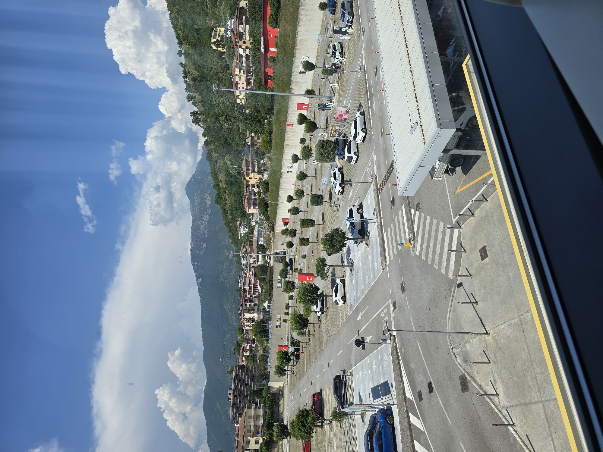 The image shows a multi-level parking area in Baronissi, Italy, on a sunny day with scattered clouds. The parking lot is paved and marked with parking spaces, including some designated for electric vehicle charging. Several cars are parked in the lot, including a blue sedan, a red hatchback, a white SUV, and a white compact car. There are also some trees and light poles scattered throughout the parking area. In the background, there are buildings, some of which appear to be residential, and a hilly, tree-covered landscape. A pedestrian crosswalk is visible in the foreground, with bollards and a yellow and white striped pole marking the crossing. A portion of a covered structure with a white roof and support pillars is visible in the bottom right of the image.