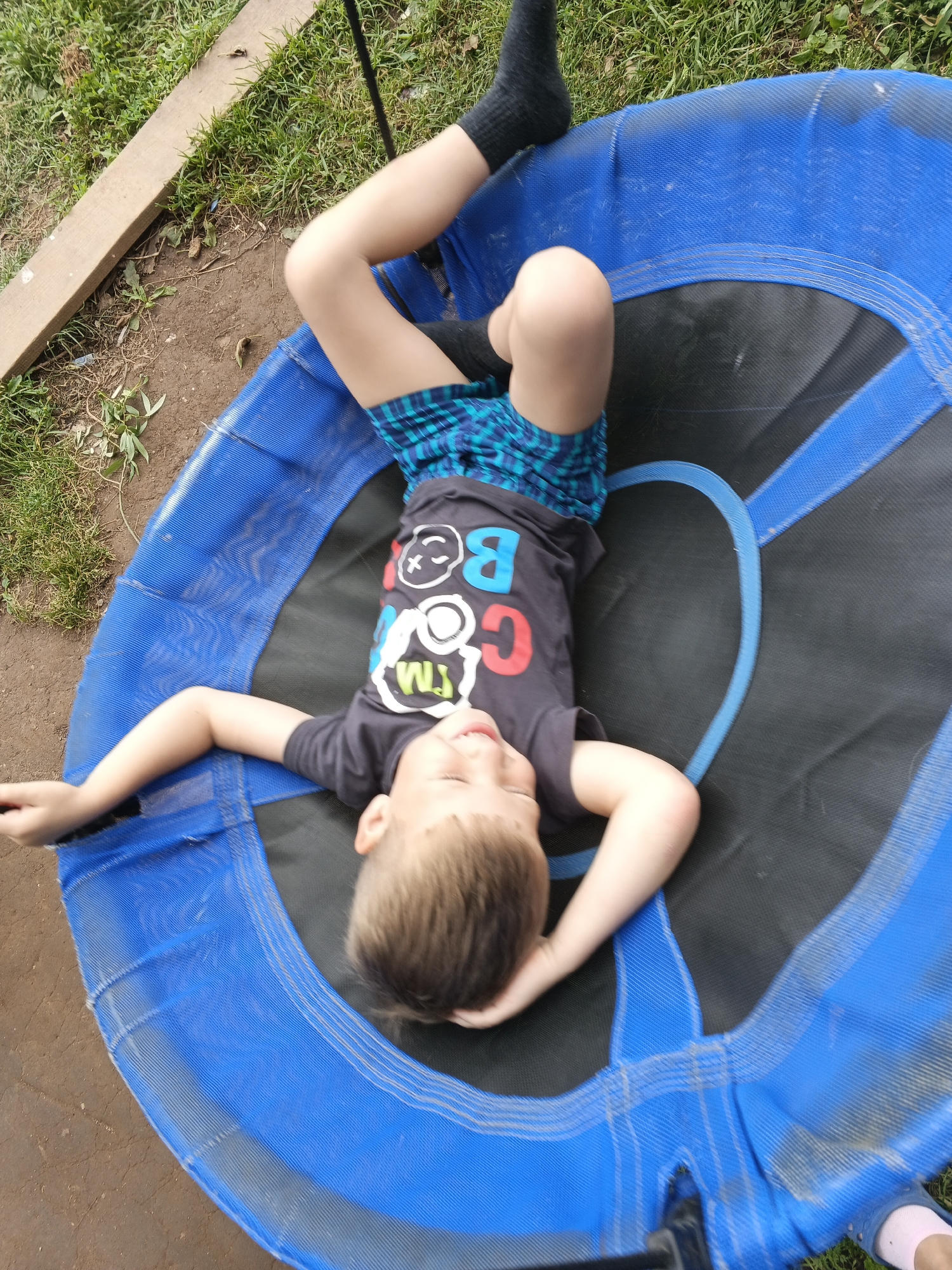A young boy lies on his back on a large, round trampoline with blue padding and a black mat. He is wearing a dark t-shirt with colorful letters that appear to spell "COOL KIDS", blue and black checkered shorts, and black socks. His arms are outstretched, with his head resting on one hand and the other arm extended to the side. He is looking upwards with a slight smile on his face. The trampoline is outdoors, with green grass and dirt visible around it. A wooden plank lies on the ground to the left of the trampoline. The lighting suggests it is daytime, and the overall scene depicts a casual, recreational moment.