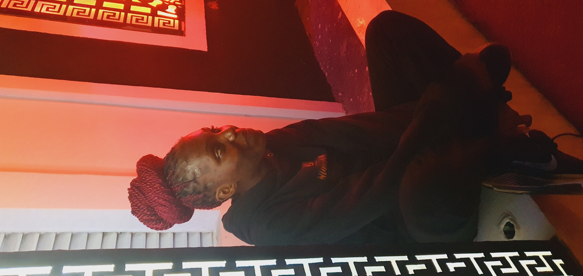 A young woman with dark skin and braided hair, styled in a large bun on top of her head, sits cross-legged on the ground. She is wearing a black hoodie and dark pants. Her head is turned to the right, and she appears to be looking at something out of frame. The background consists of a white wall with vertical blinds on the left, and a black wall with a decorative pattern on the right. The scene is illuminated by warm, reddish-orange lighting, casting shadows on the ground. There is a portion of a white structure or ledge in the foreground where the woman is sitting. The environment suggests an outdoor or semi-outdoor setting, possibly at night or in a dimly lit area.