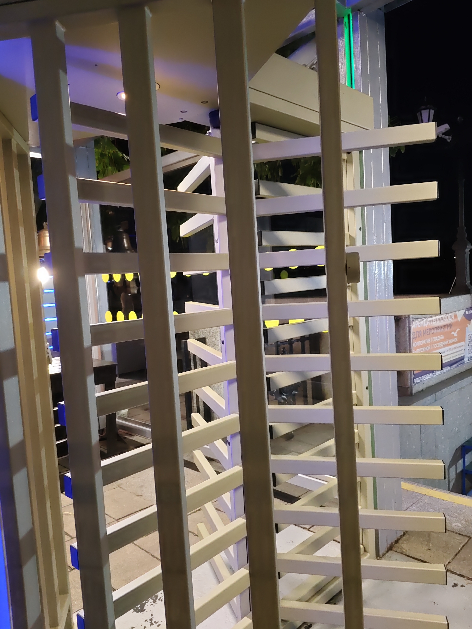 The image shows a turnstile, likely an entrance to a paid area, located outdoors at night. The turnstile is made of light-colored metal bars. In the background, there are trees and some lighting. A sign is visible on the right side of the image with Russian text, which appears to be an advertisement for renting venues for events. The text on the sign includes "АРЕНДА ТЕПЛОХОДА", "ДЛЯ МЕРОПРИЯТИЙ", and other phrases related to corporate events, weddings, and celebrations. The general environment suggests an urban setting, possibly a park or an entertainment venue. The city of Rostov-on-Don, Russia is identified as the location.