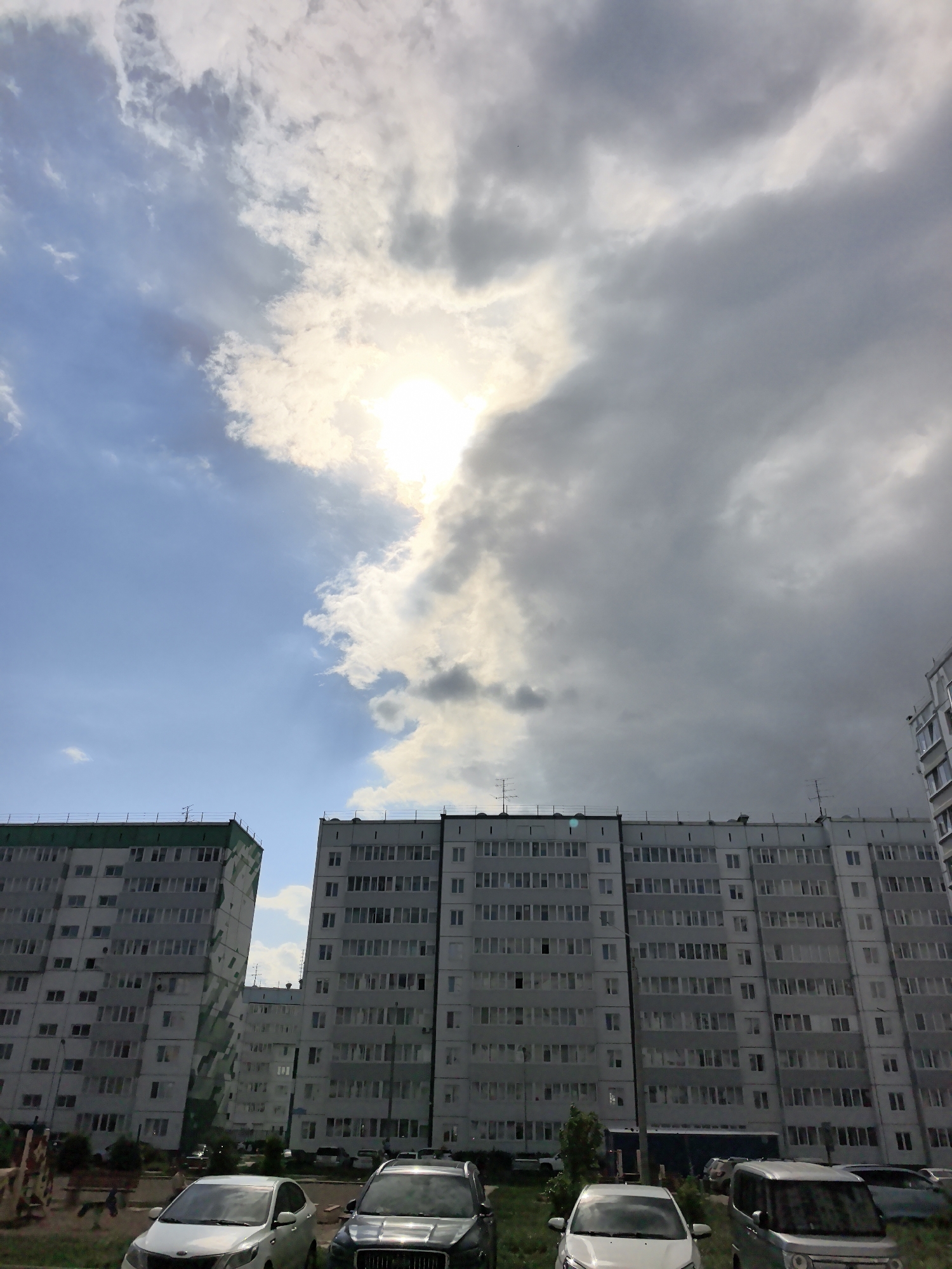 The image depicts a cityscape in Фролы, Russia, with a focus on two multi-story apartment buildings under a partly cloudy sky. The building on the left has a green-trimmed roof, while the building on the right is plain. A parking lot is visible in the foreground with several parked cars. The sky is divided with a blue area and the sun shining, while dark clouds suggesting possible rain.