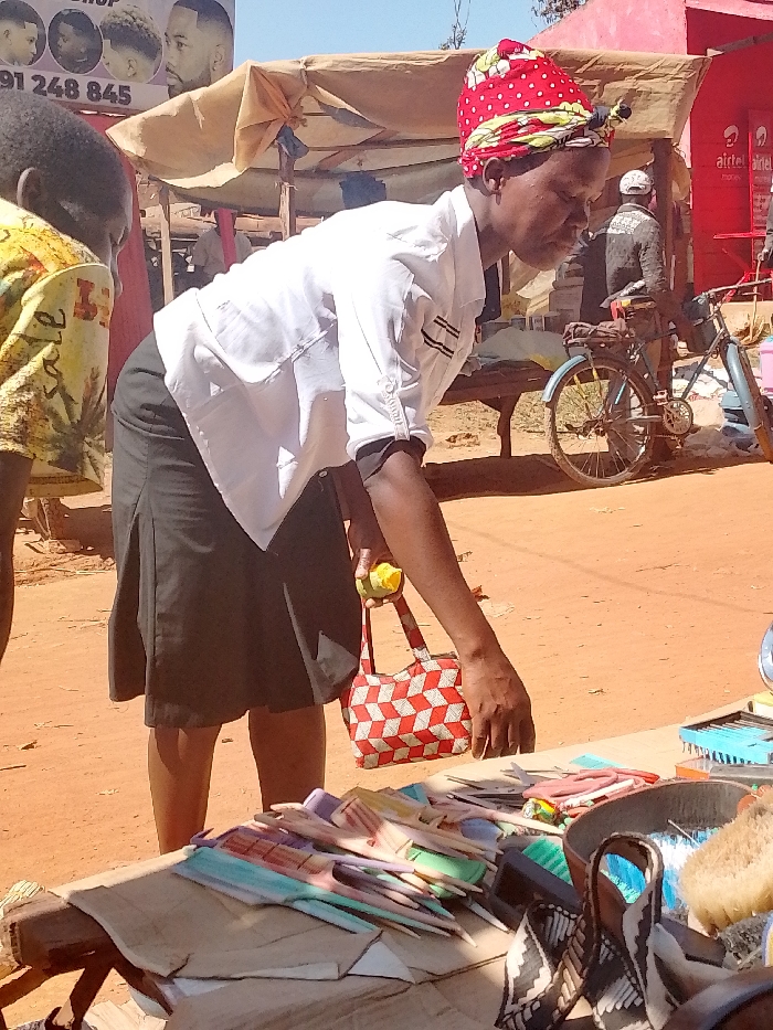 The image depicts a woman, wearing a white shirt and black skirt, holding a red and white checkered purse, and a small yellow fruit, standing near a table covered in assorted combs and hairbrushes. The woman is looking down at the table, possibly selecting a comb or brush to purchase. Behind the table, a bicycle sits parked nearby, with a red building in the background. The scene takes place in a marketplace in Lwandeti, Kenya, near the location with coordinates (latitude: 0.563, longitude: 34.806). The setting appears to be a bustling, open-air market with various vendors selling their wares. The woman is the main subject of the image, while the table of combs and brushes and the bicycle are secondary subjects. The overall atmosphere of the scene is vibrant and active. The image appears to have been taken in the daytime, with sunlight illuminating the scene. The ground is a mix of dirt and sand, typical of a market setting in Lwandeti, Kenya. The overall scene captures the everyday hustle and bustle of life in Lwandeti, Kenya, with a focus on the woman's transaction at the market.