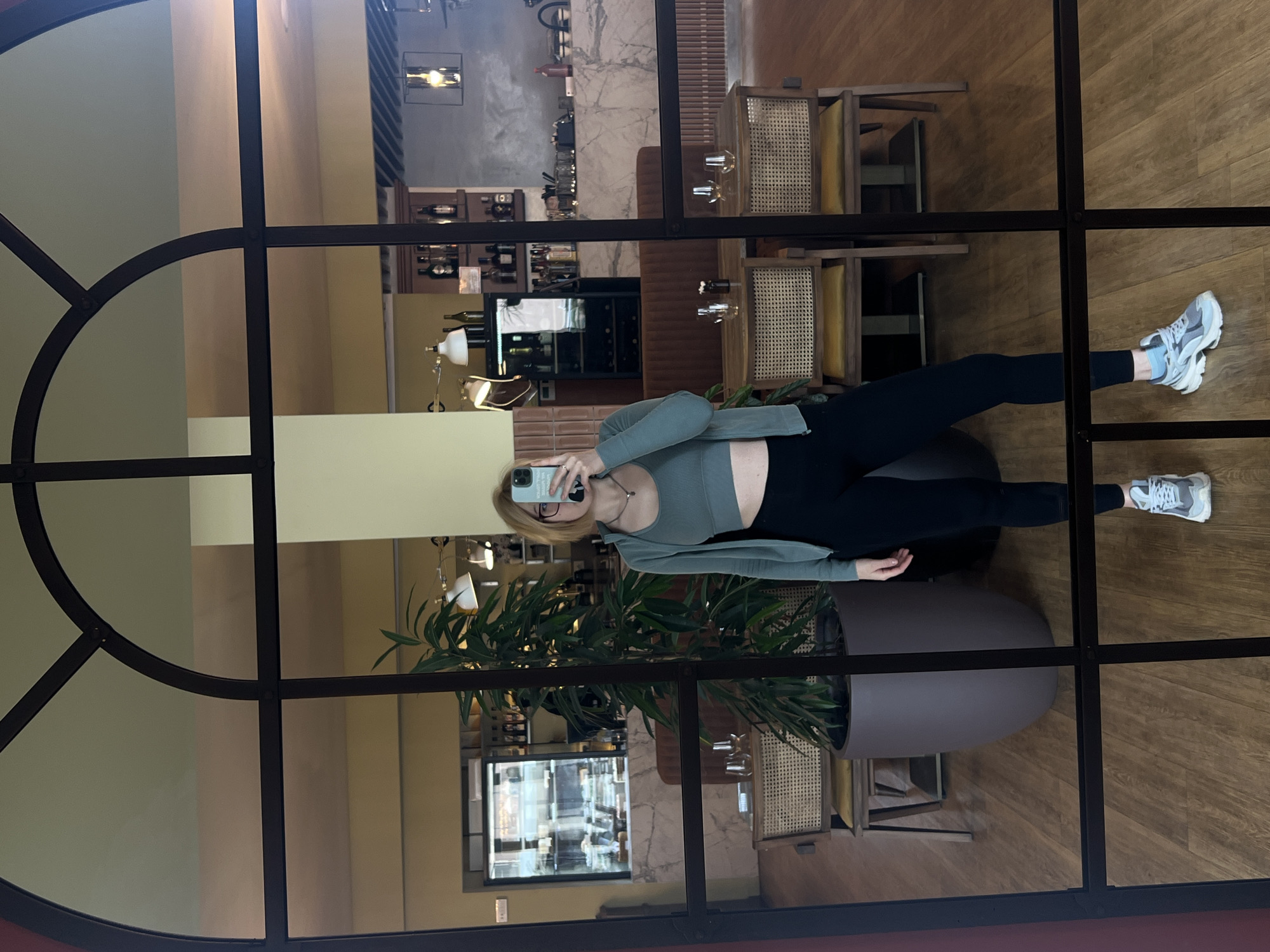 A woman in athletic attire (green top, black leggings, gray running shoes) is taking a selfie in a mirror with a complex frame, likely indoors. The reflection shows a restaurant interior with tables, chairs, a plant in a large pot, and a wine rack. The location appears to be a restaurant setting based on the furnishings and reflection in the glass, possibly located in or around Boreti, Montenegro, judging by the architecture and style of the interior. The woman is holding a phone up to take a picture of herself in the mirror.