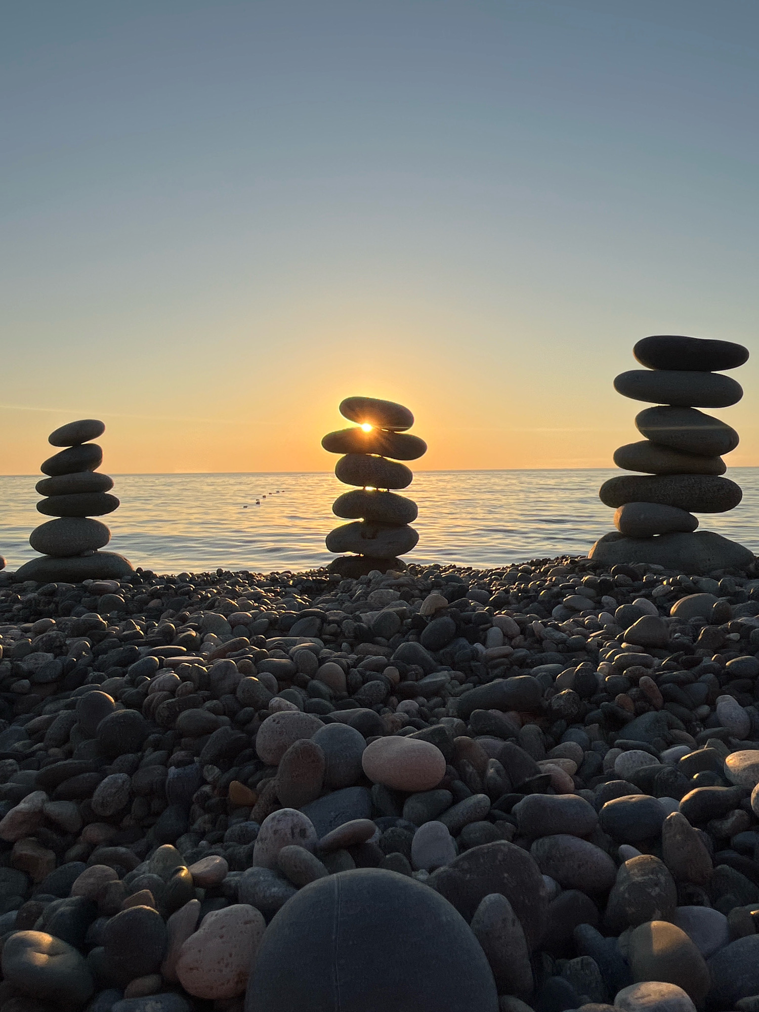 The image depicts a serene sunset scene on the pebble beach of Kobuleti, Georgia. Three meticulously stacked cairns of various sizes are positioned on the shore, with the largest one slightly to the left, a smaller one in the center with the sun peeking through its stones, and another medium-sized cairn to the right. The calm sea stretches out towards the horizon, reflecting the warm hues of the setting sun. The sky above transitions from a soft blue to a warm orange glow near the horizon, indicating the golden hour. The foreground is filled with a multitude of smooth, dark pebbles, characteristic of Kobuleti's beaches. The overall impression is one of tranquility and natural beauty, with the stacked stones adding an element of human intervention and artistic arrangement to the coastal landscape.