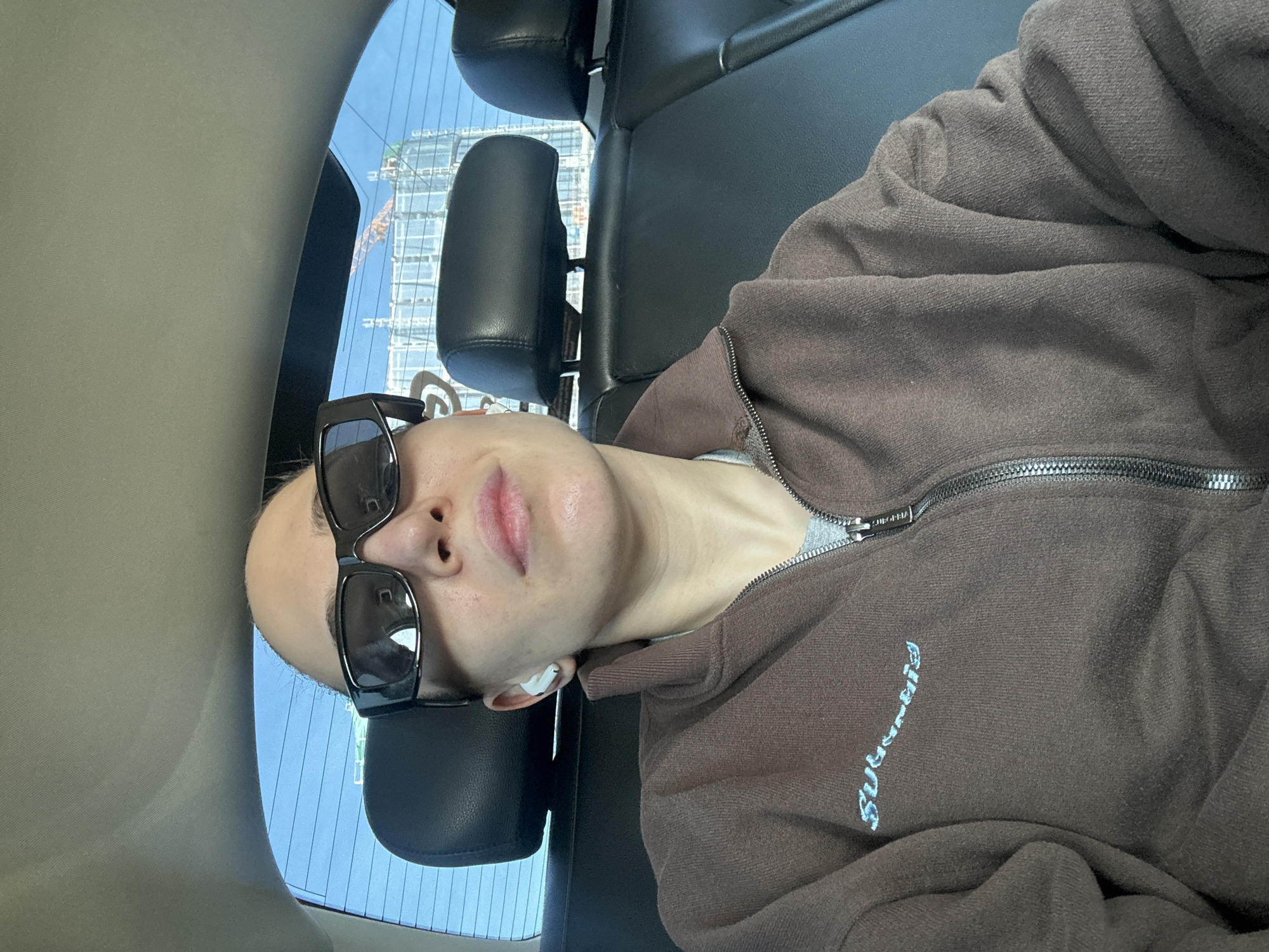 A person with a shaved head is sitting in the back seat of a car, wearing sunglasses and an earbud. The person is wearing a brown zip-up jacket with blue lettering on the front. Outside the car window, a building under construction with a crane is visible, indicating a possible location in a rapidly developing area of Moscow. The car has black leather seats and a light grey ceiling.
